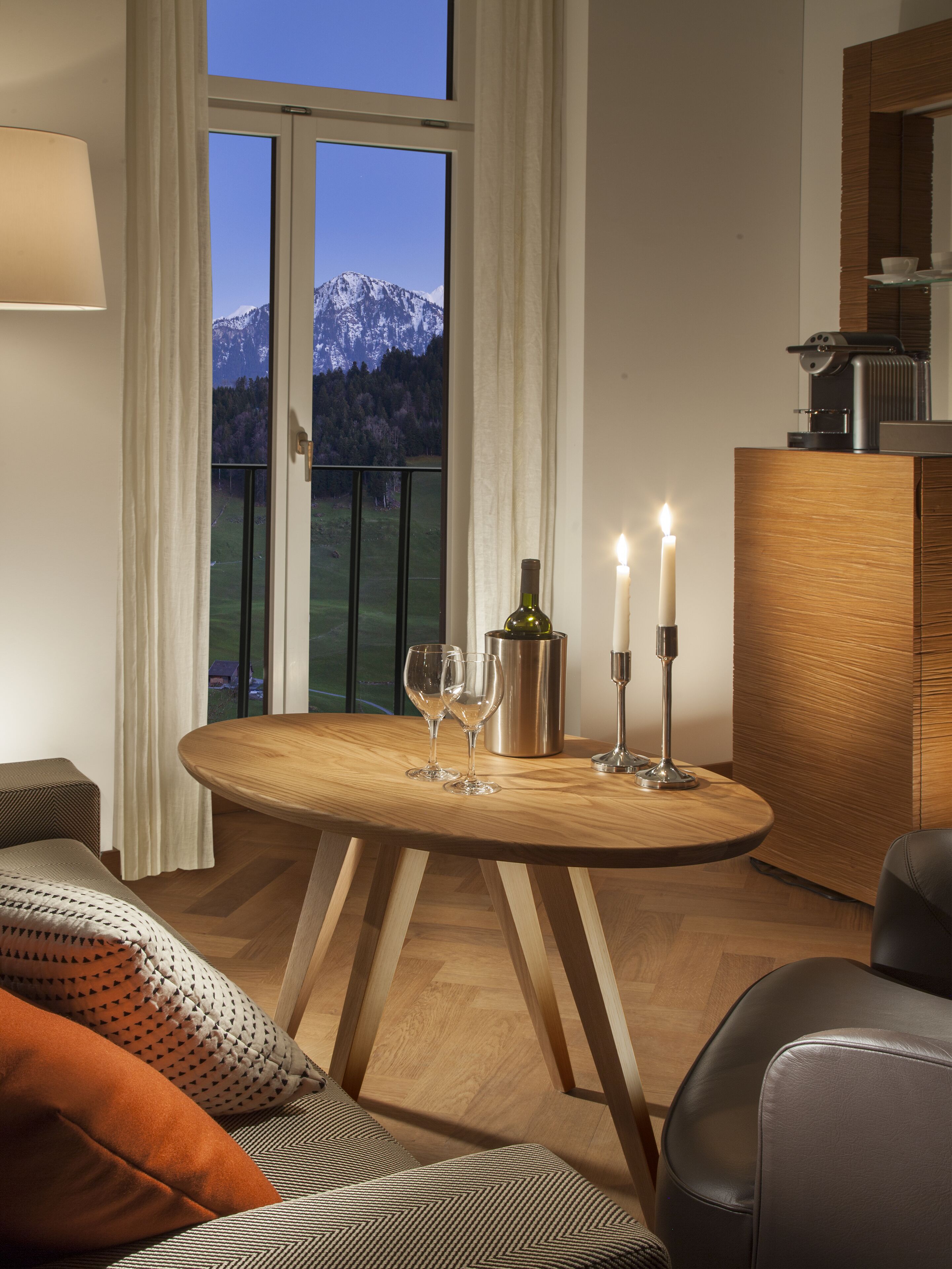 A dining area with a wooden table, two wine glasses, a candle, and a bottle. A view of mountains is visible through the window.