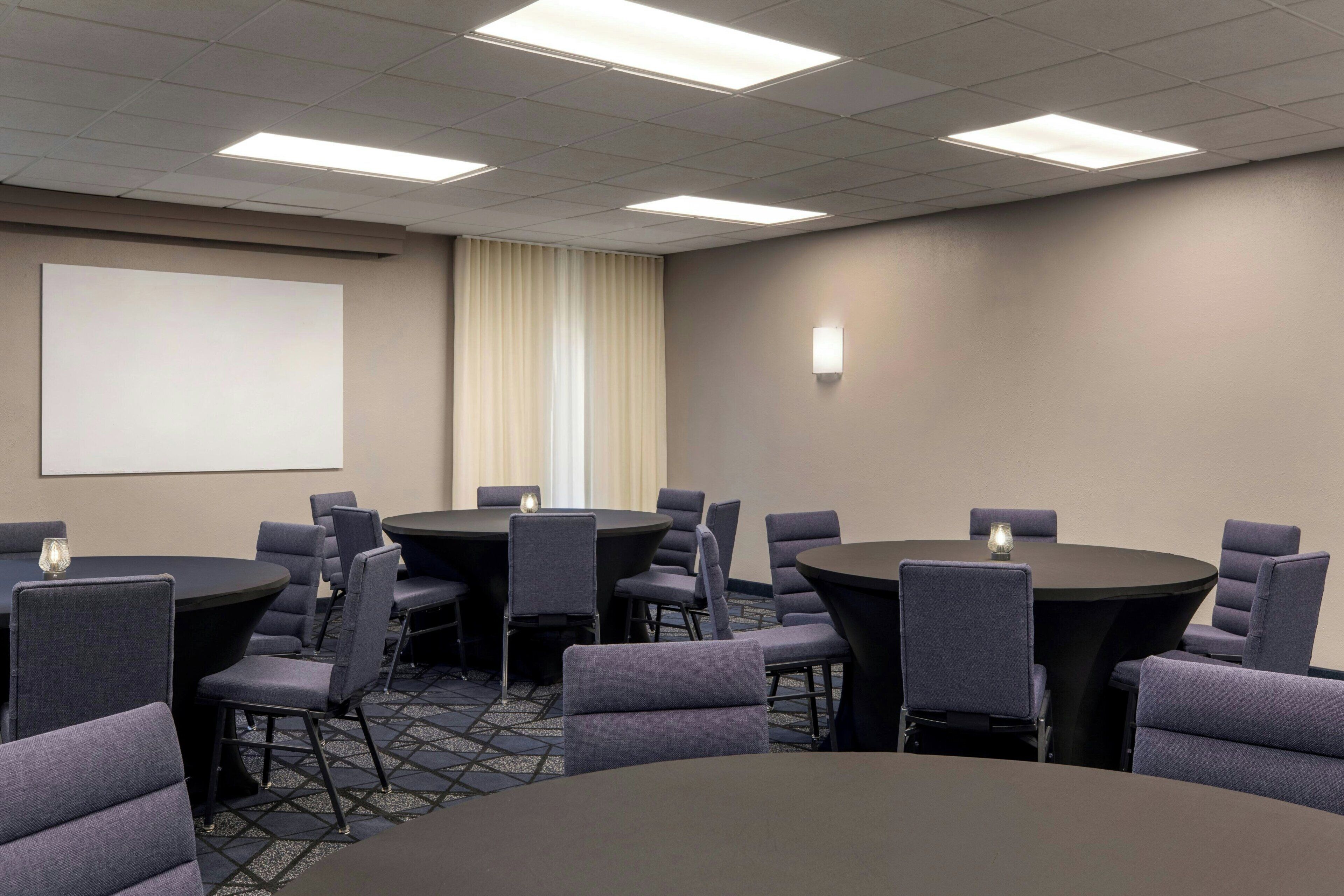 A conference room with multiple tables and chairs, a projector screen, and a large window with curtains.