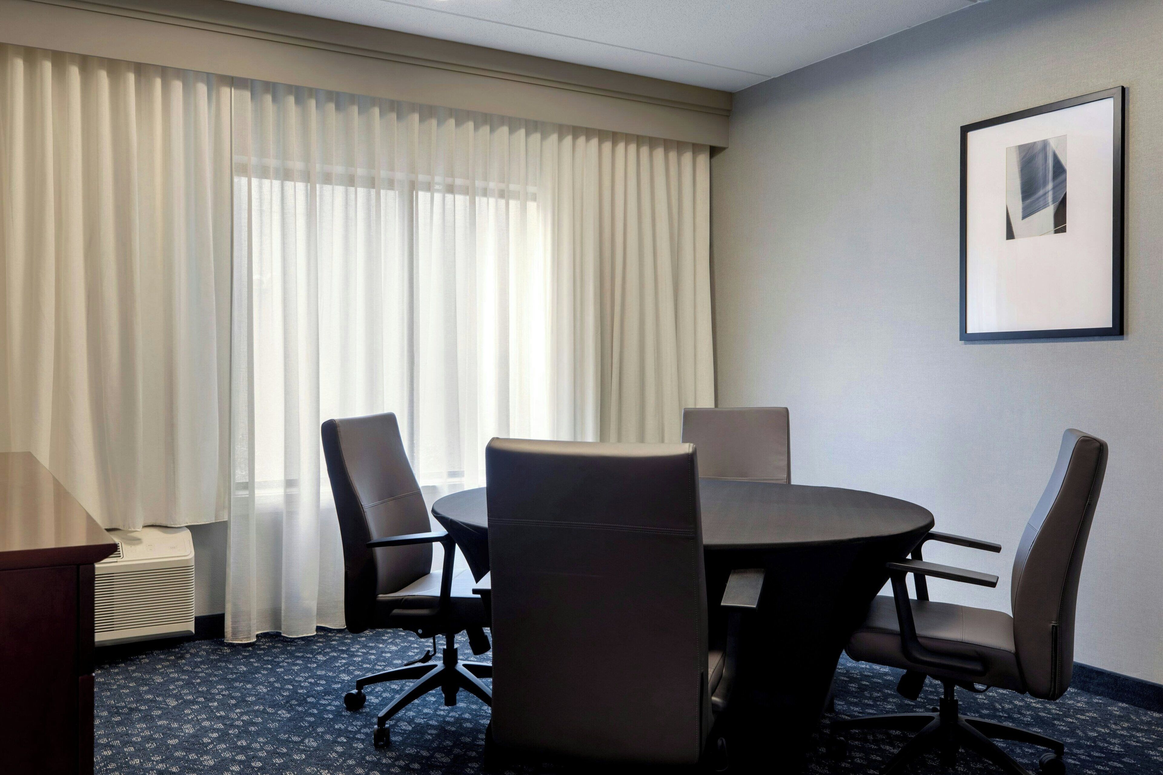 A conference room with a round table, four chairs, a framed picture on the wall, and a window with curtains.