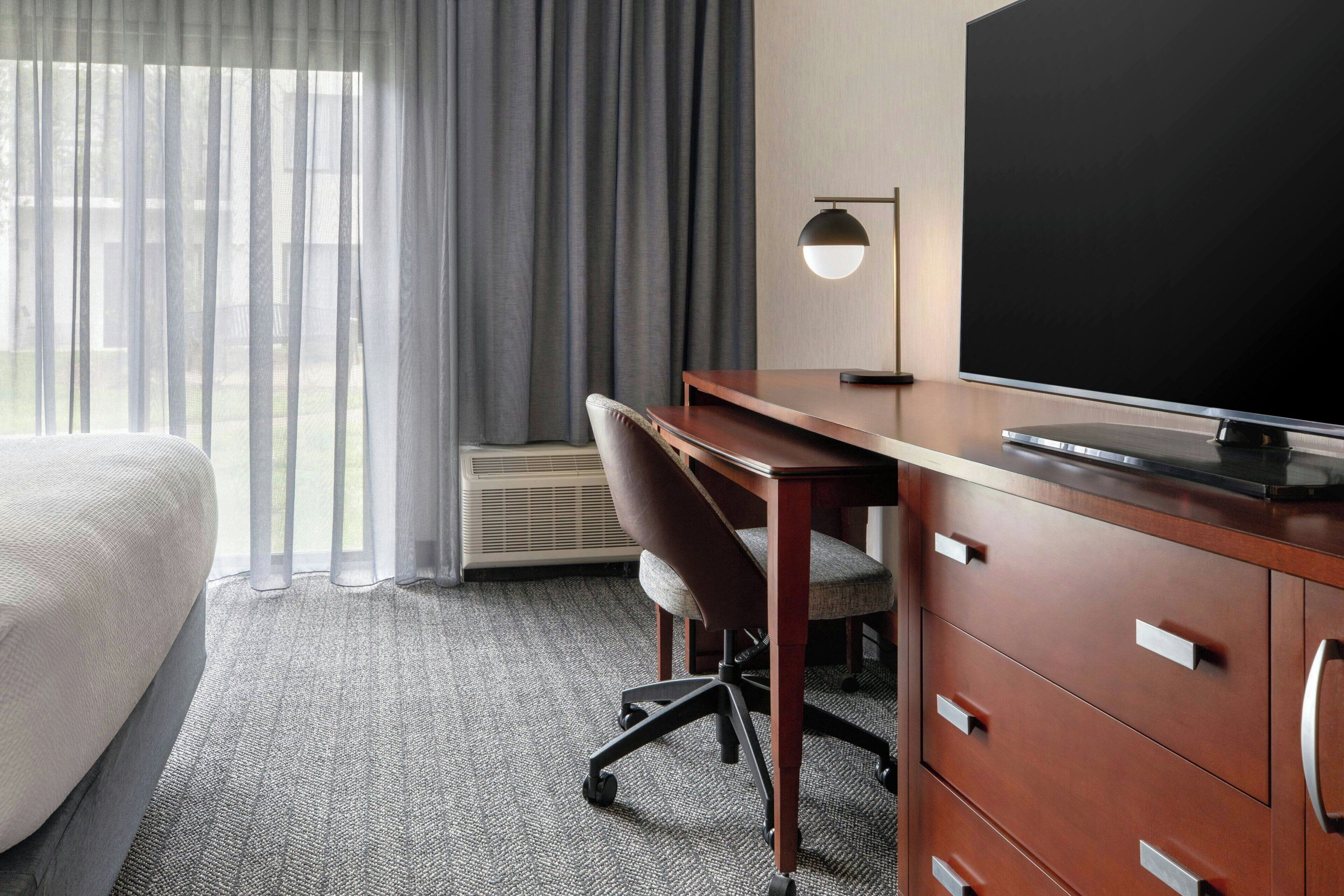 Habitación de hotel con cama, escritorio con silla, televisor de pantalla plana, lámpara y ventana grande con cortinas.