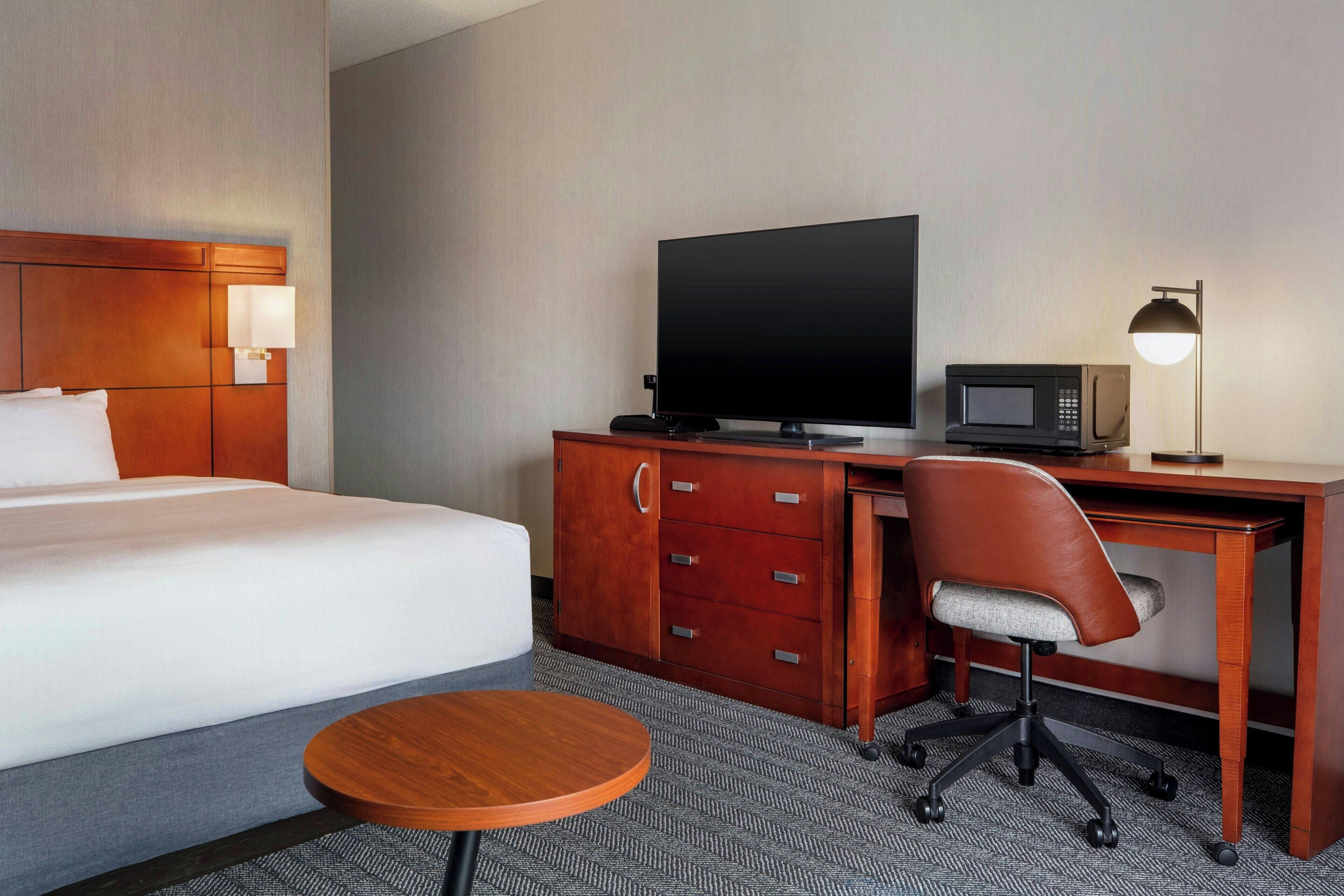 A hotel room with a bed, a desk with a television, a microwave, a chair, and a small round table.