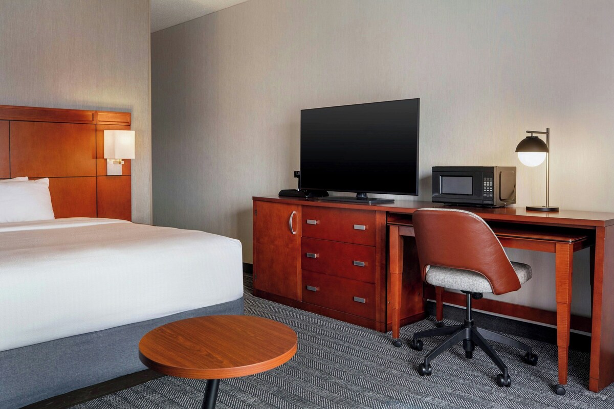 A hotel room with a bed, a desk with a television, a microwave, a chair, and a small round table.