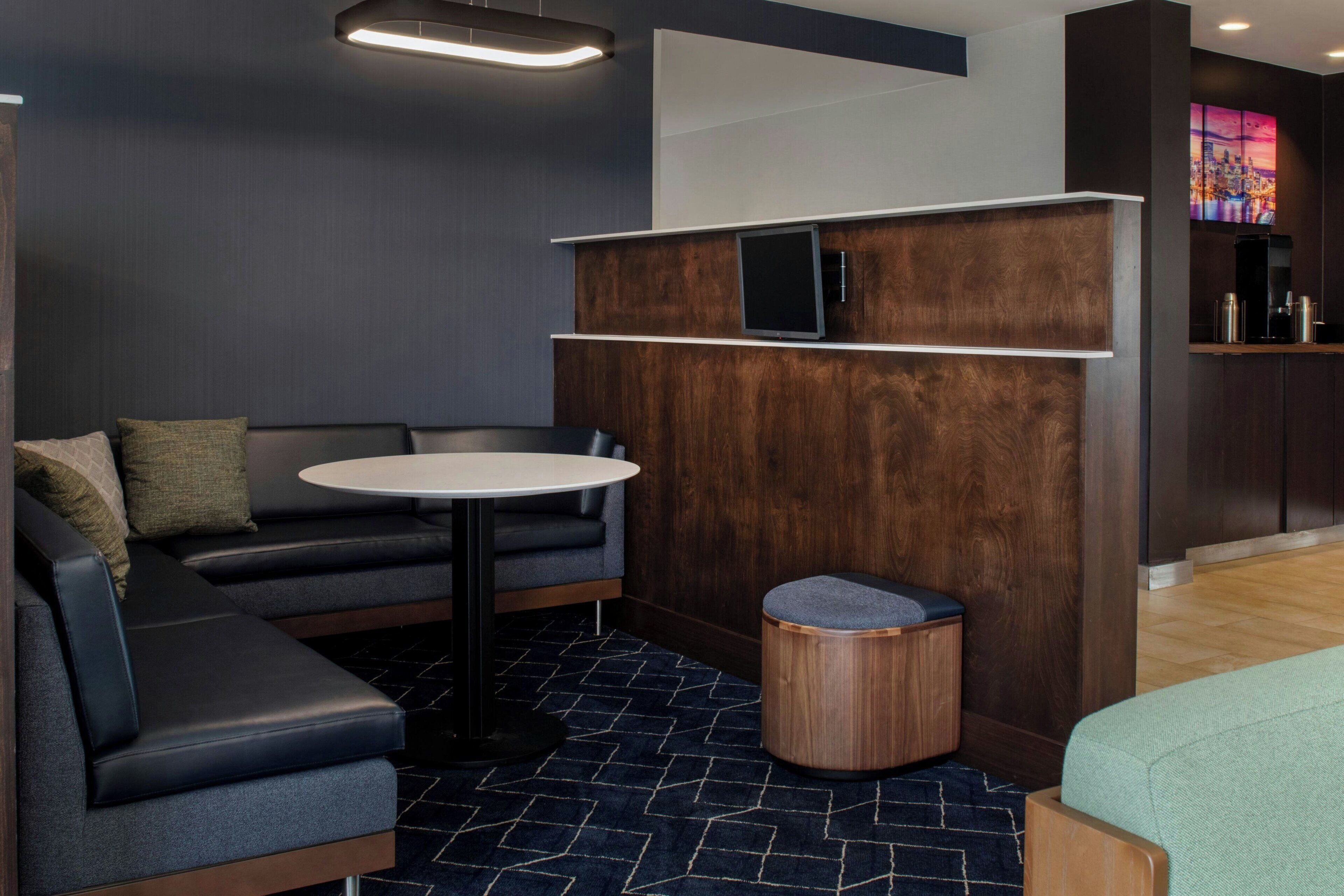 Une chambre d’hôtel moderne, dotée d’un canapé d’angle, d’une table ronde, d’un petit tabouret et d’un téléviseur à écran plat fixé sur un panneau en bois.