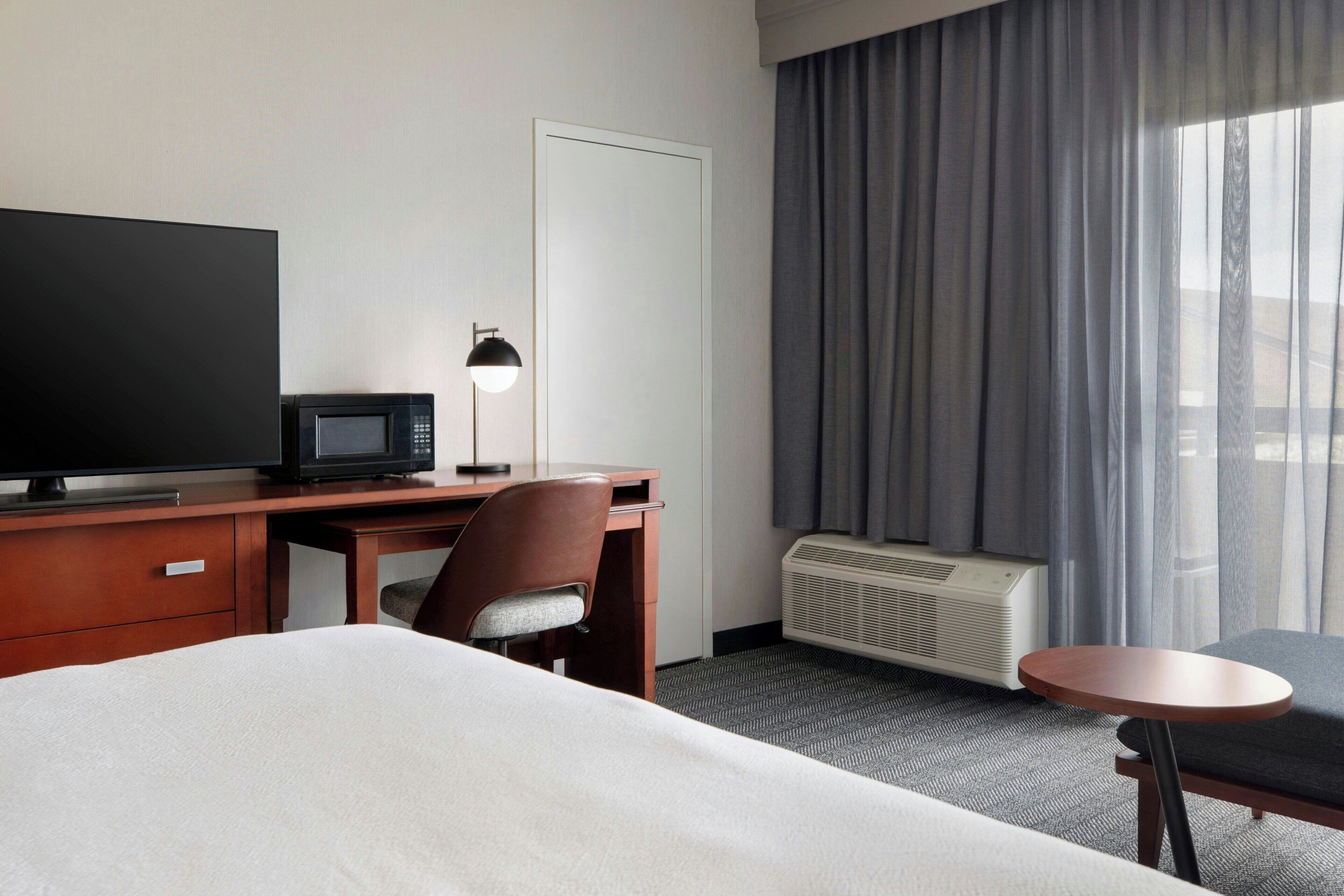 Habitación de hotel con cama, escritorio con televisor y lámpara, una silla, una mesita y ventana con cortinas.