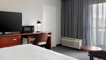 Habitación de hotel con cama, escritorio con televisor y lámpara, una silla, una mesita y ventana con cortinas.