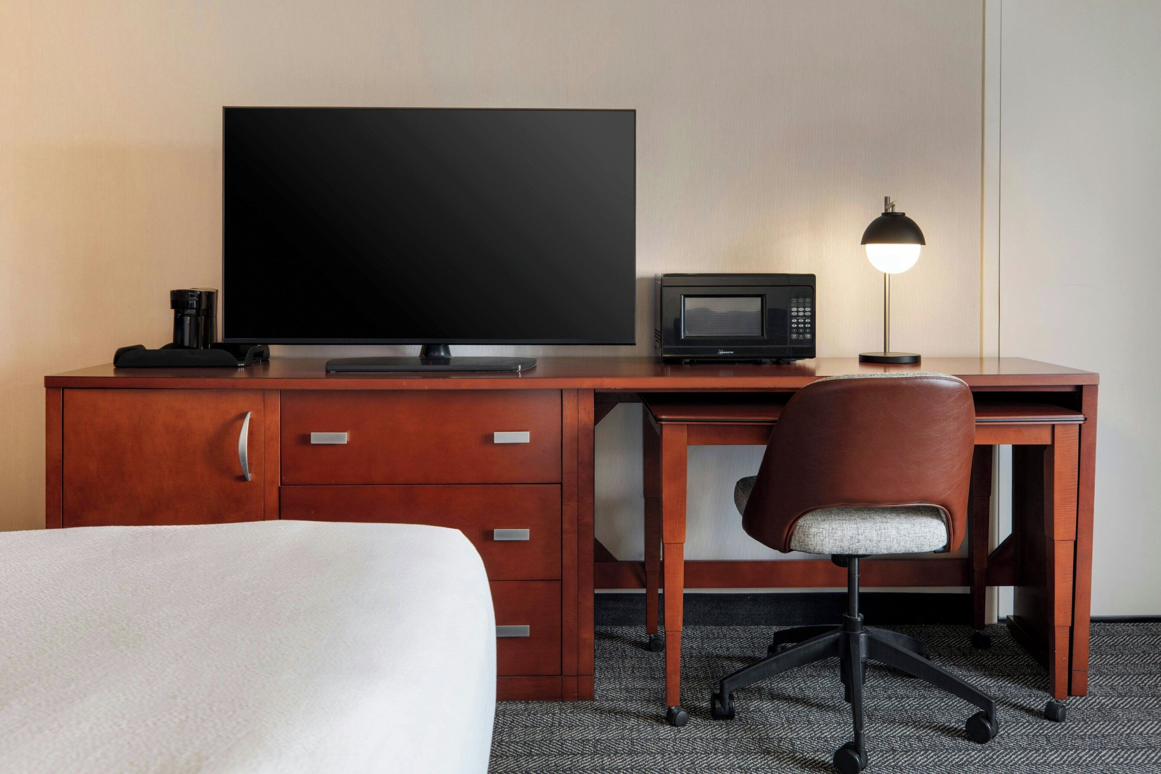 A hotel room with a bed, a wooden desk with a flat-screen TV, a microwave, a lamp, and a brown office chair.