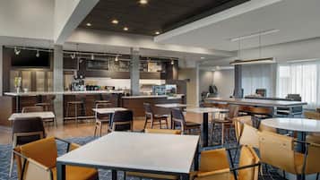 A modern hotel lobby with a bar area, seating arrangements, and a visible kitchen in the background.