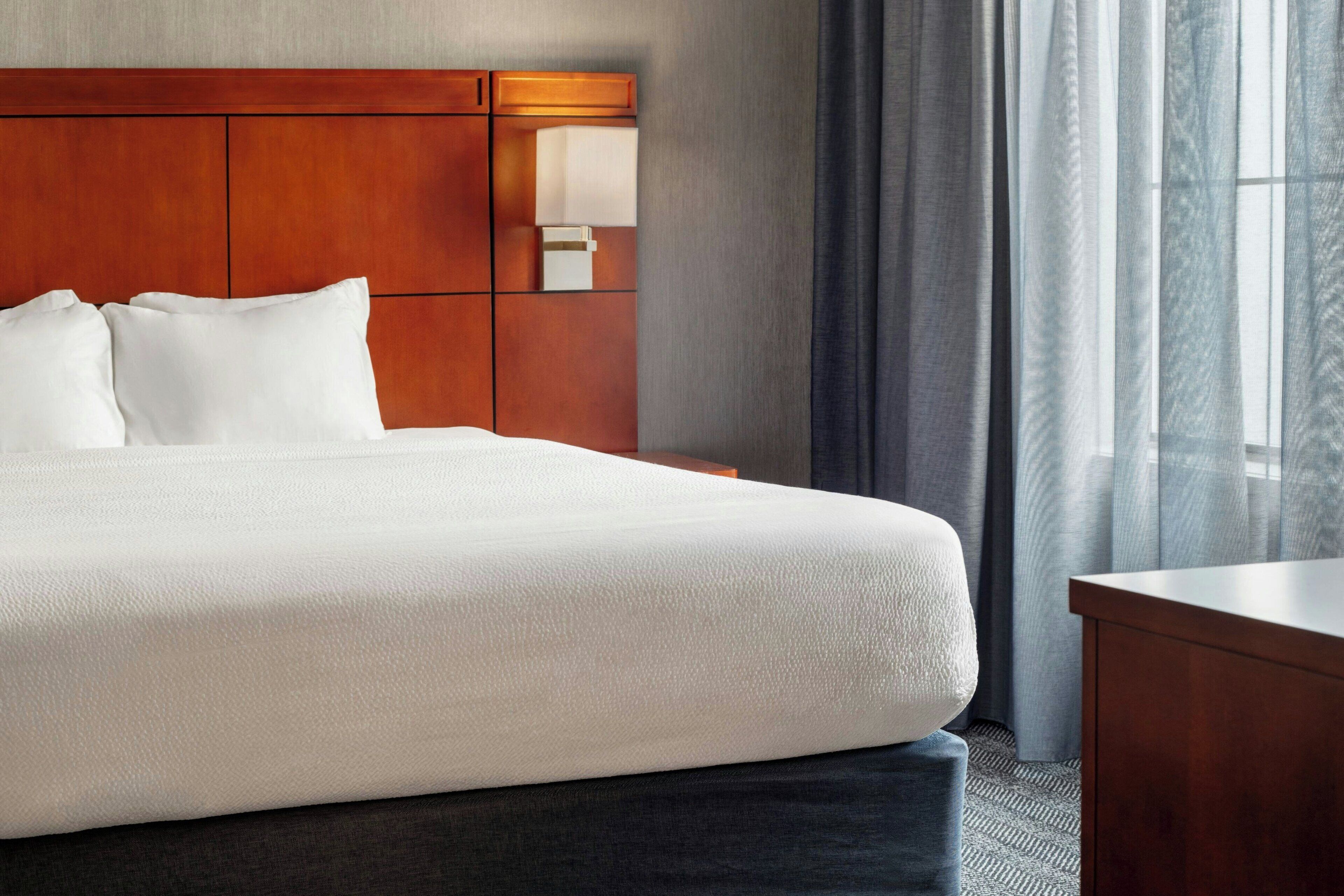 A hotel room with a large bed, a wooden headboard, a nightstand, a lamp, and a view of the outdoors through sheer curtains.