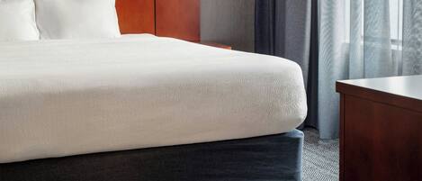A hotel room with a large bed, a wooden headboard, a nightstand, a lamp, and a view of the outdoors through sheer curtains.