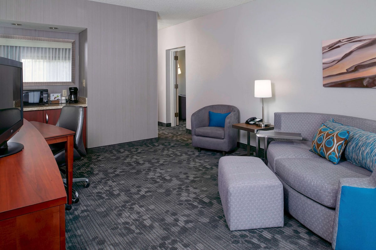 Une chambre d’hôtel avec un bureau, un canapé gris, un fauteuil gris, une petite table avec une lampe et un tableau au mur.