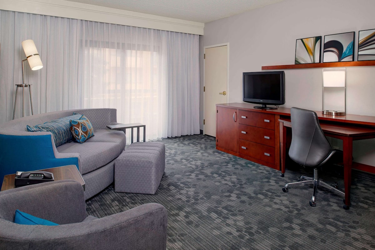 Une chambre d’hôtel avec un canapé gris, un petit pouf, un bureau avec une chaise, une télévision et un meuble.