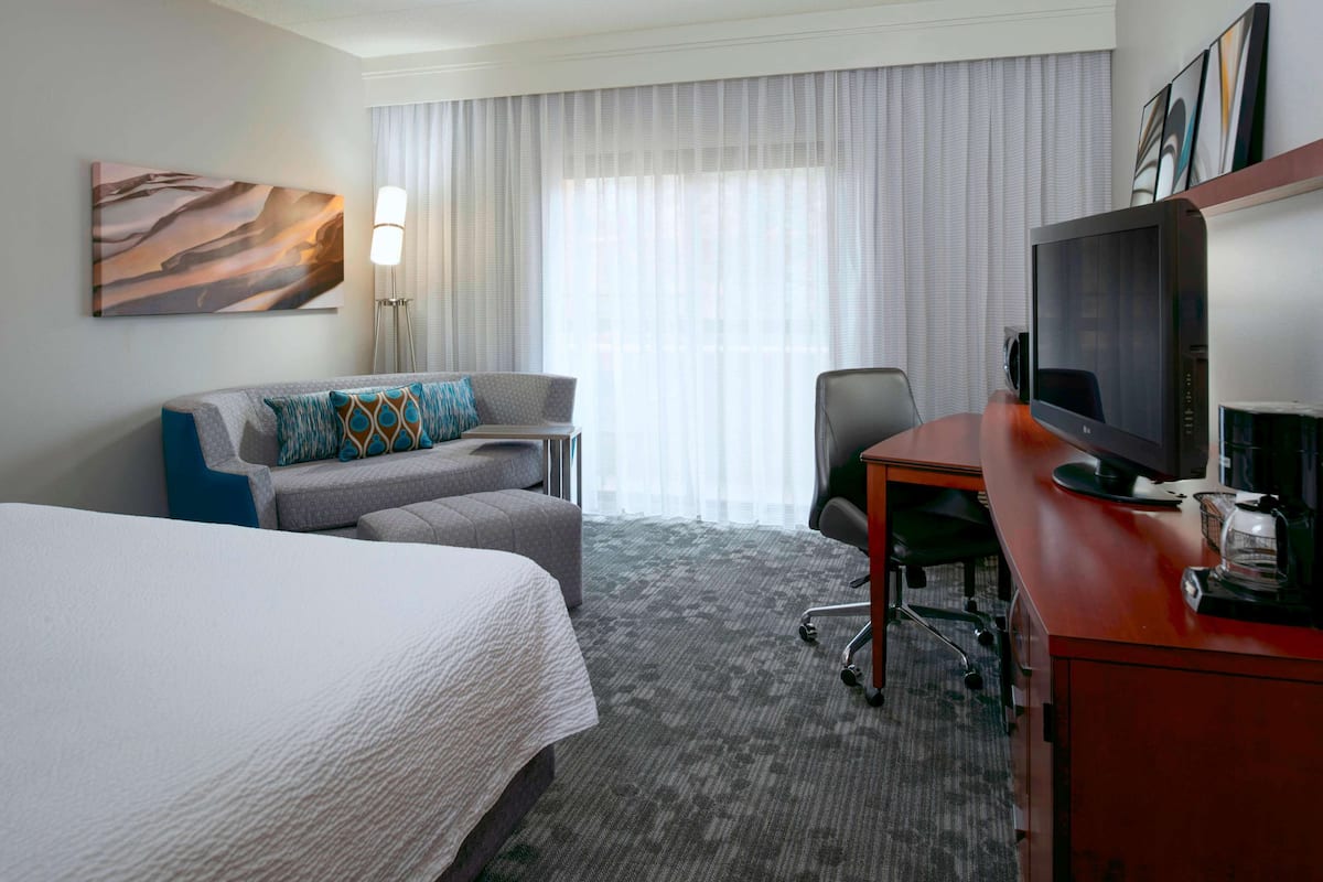 Une chambre d’hôtel avec un lit, un bureau avec un ordinateur, une chaise, un canapé, une lampe et une grande fenêtre avec des rideaux.
