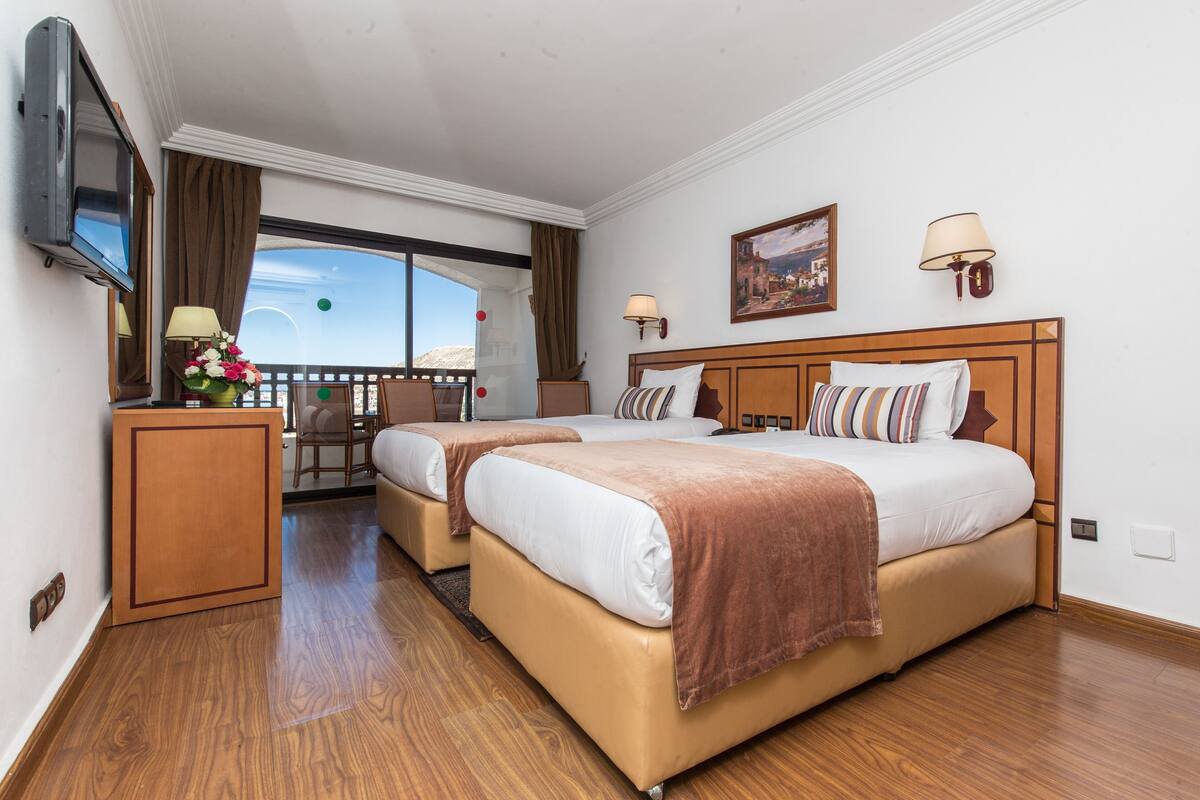 A hotel room with two beds, a TV, a balcony with a view, and a wooden floor.