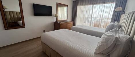 Habitación de hotel con dos camas, televisión, espejo, cómoda y ventana con cortinas.