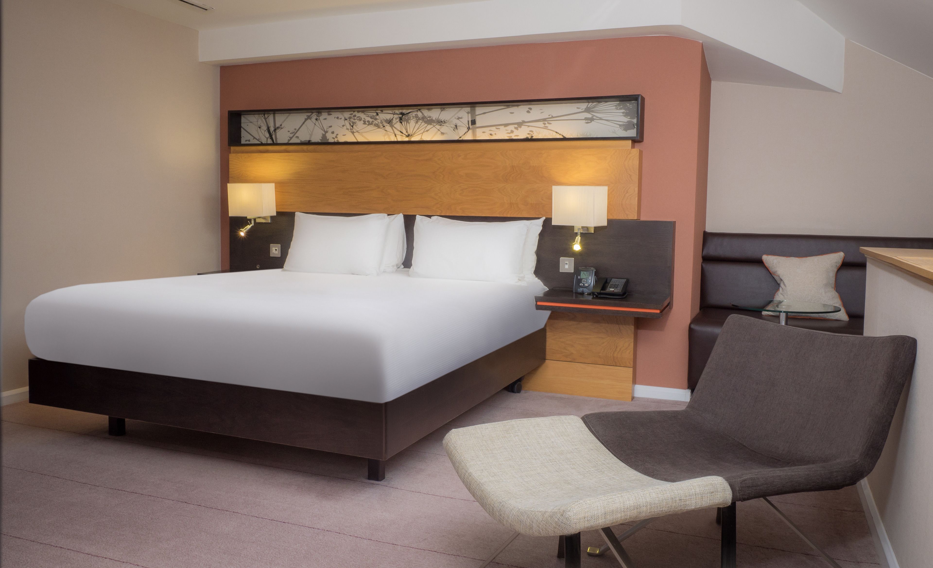 A hotel room with a large bed, a bedside table with a lamp, a chair, and a small desk with a phone.