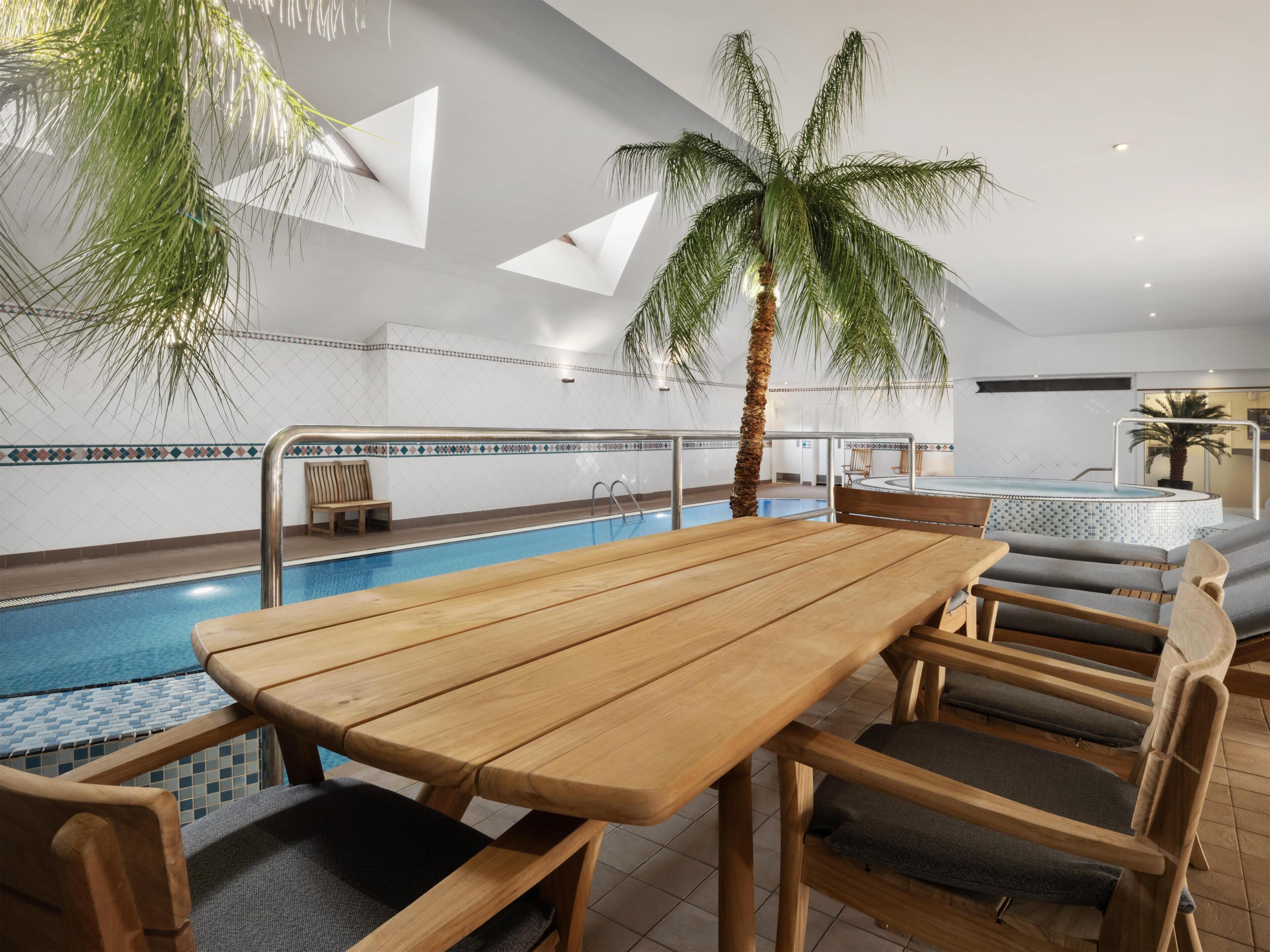 A poolside area with a wooden table and chairs, a palm tree, and a tiled pool.