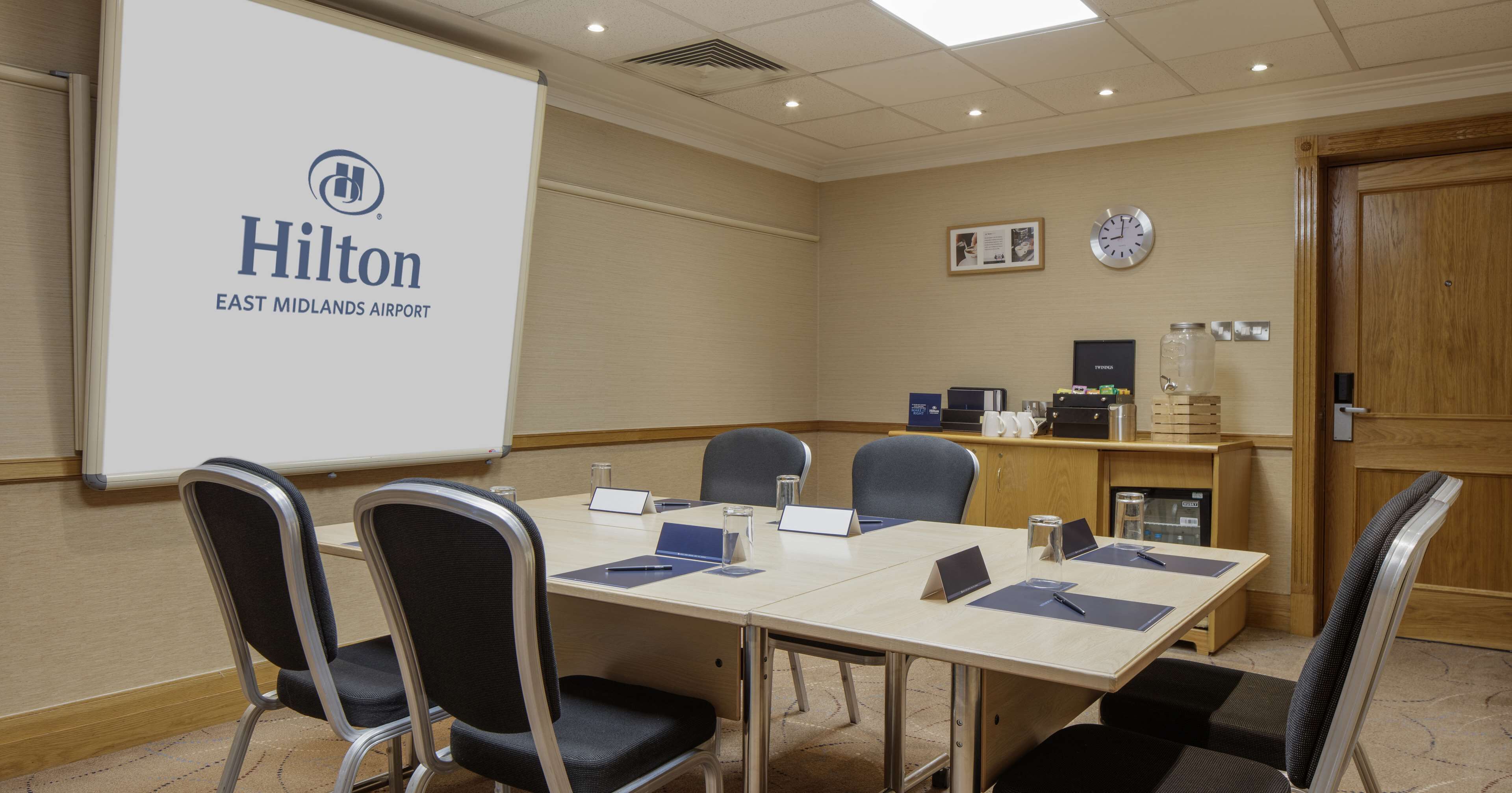 Une salle de conférence dotée d’un grand écran affichant le logo de Hilton, d’un bureau en bois et de plusieurs chaises.