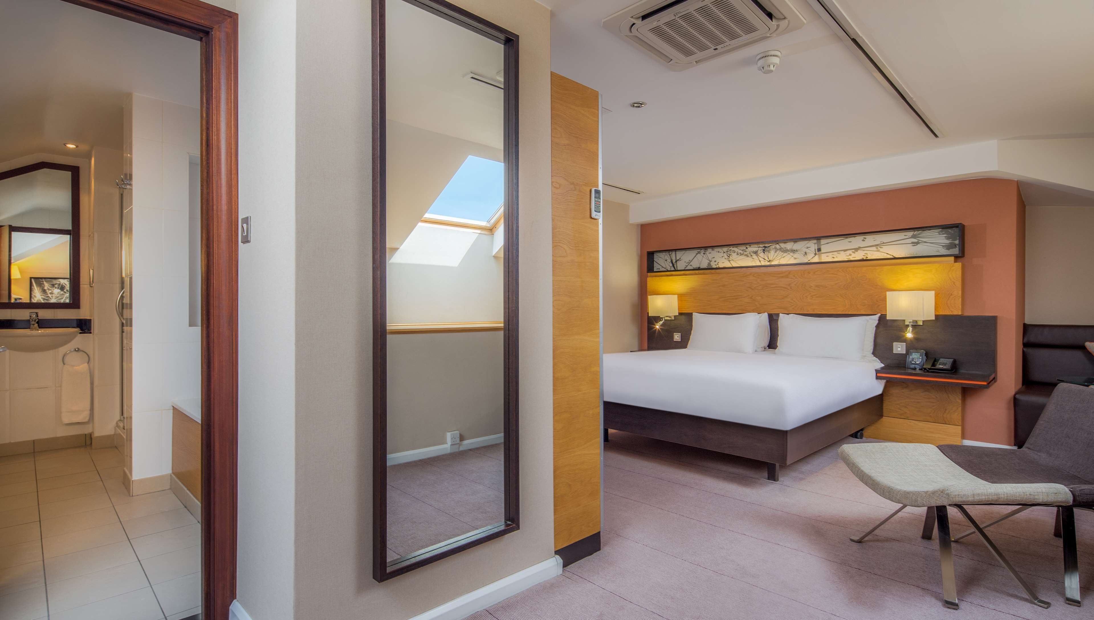 A hotel room with a large bed, bedside tables with lamps, a chair, a mirror, and a skylight.
