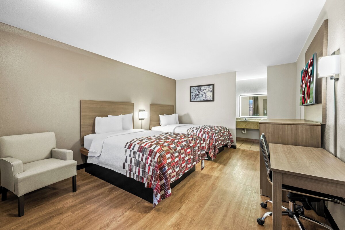 A hotel room with two beds, a desk, a chair, a sofa, and a bathroom visible through an open door.