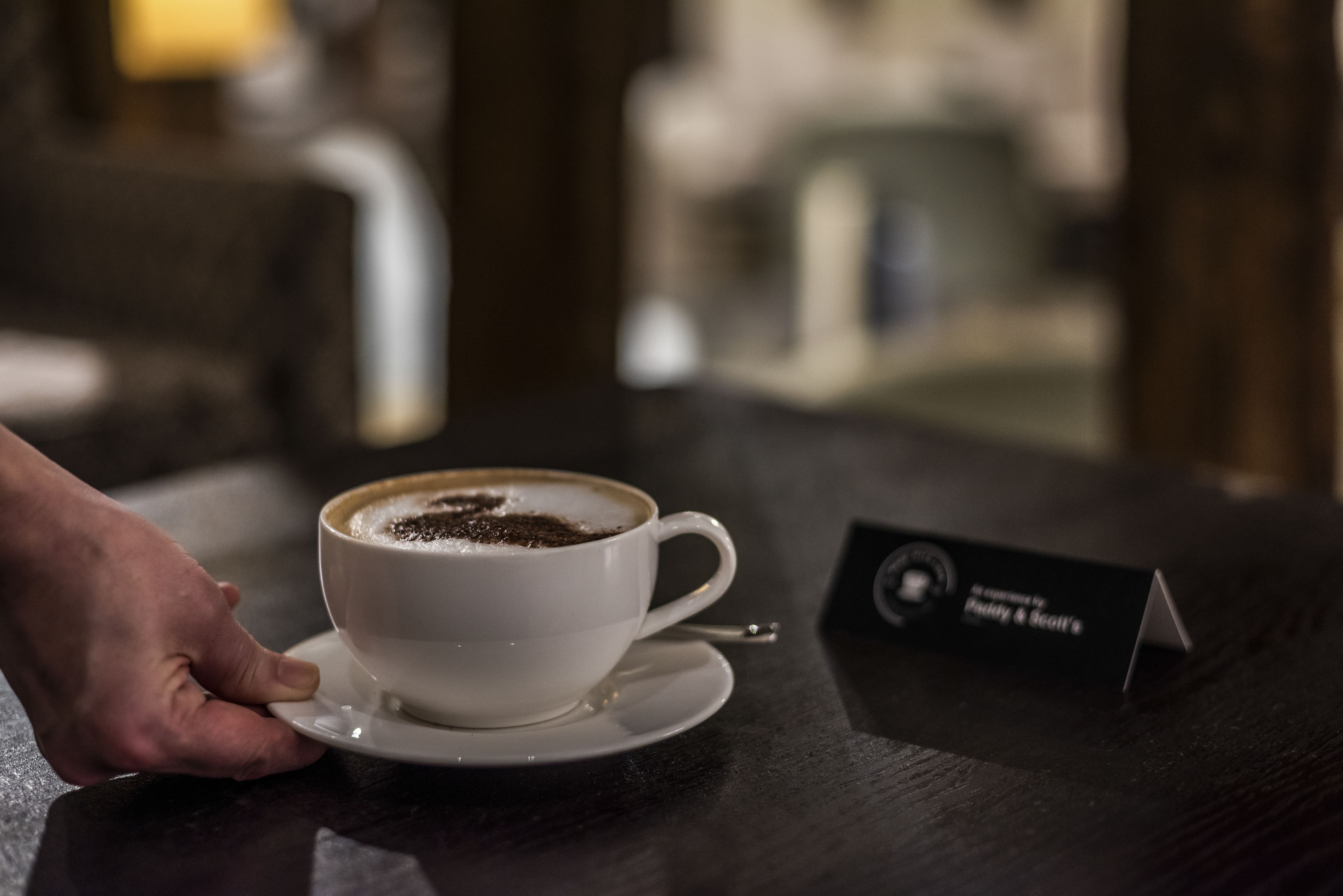 Una taza de café sobre un platito, una mano sosteniendo el platito, y una tarjeta de presentación sobre la mesa.