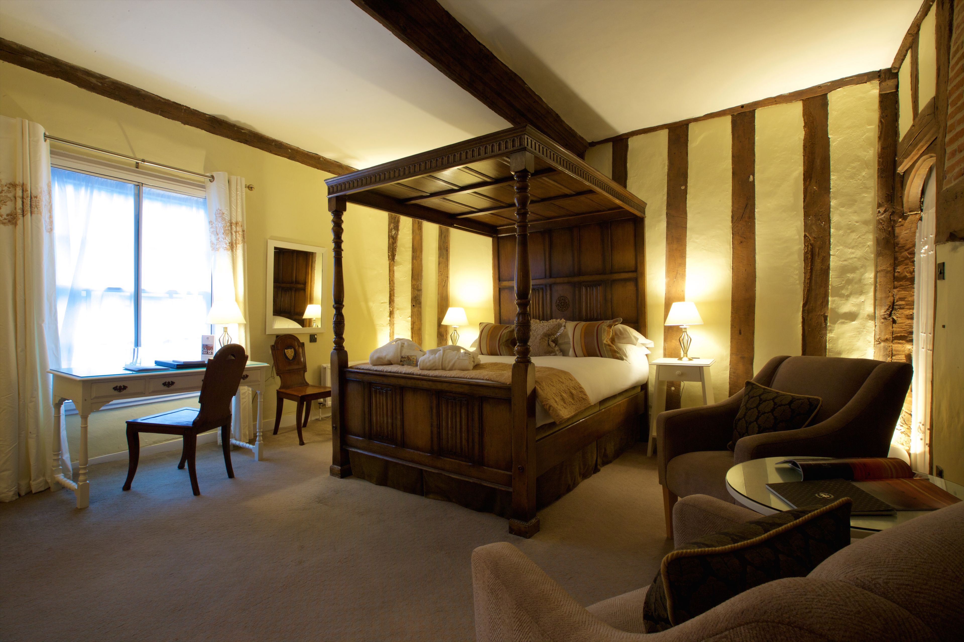 A traditional four-poster bed with a wooden headboard, a desk with a chair, a sofa, and a small table with a lamp.