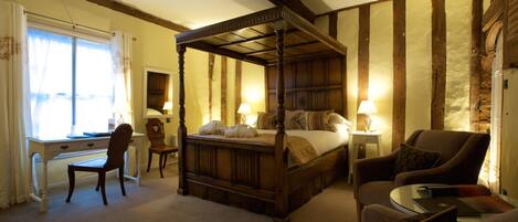 A traditional four-poster bed with a wooden headboard, a desk with a chair, a sofa, and a small table with a lamp.