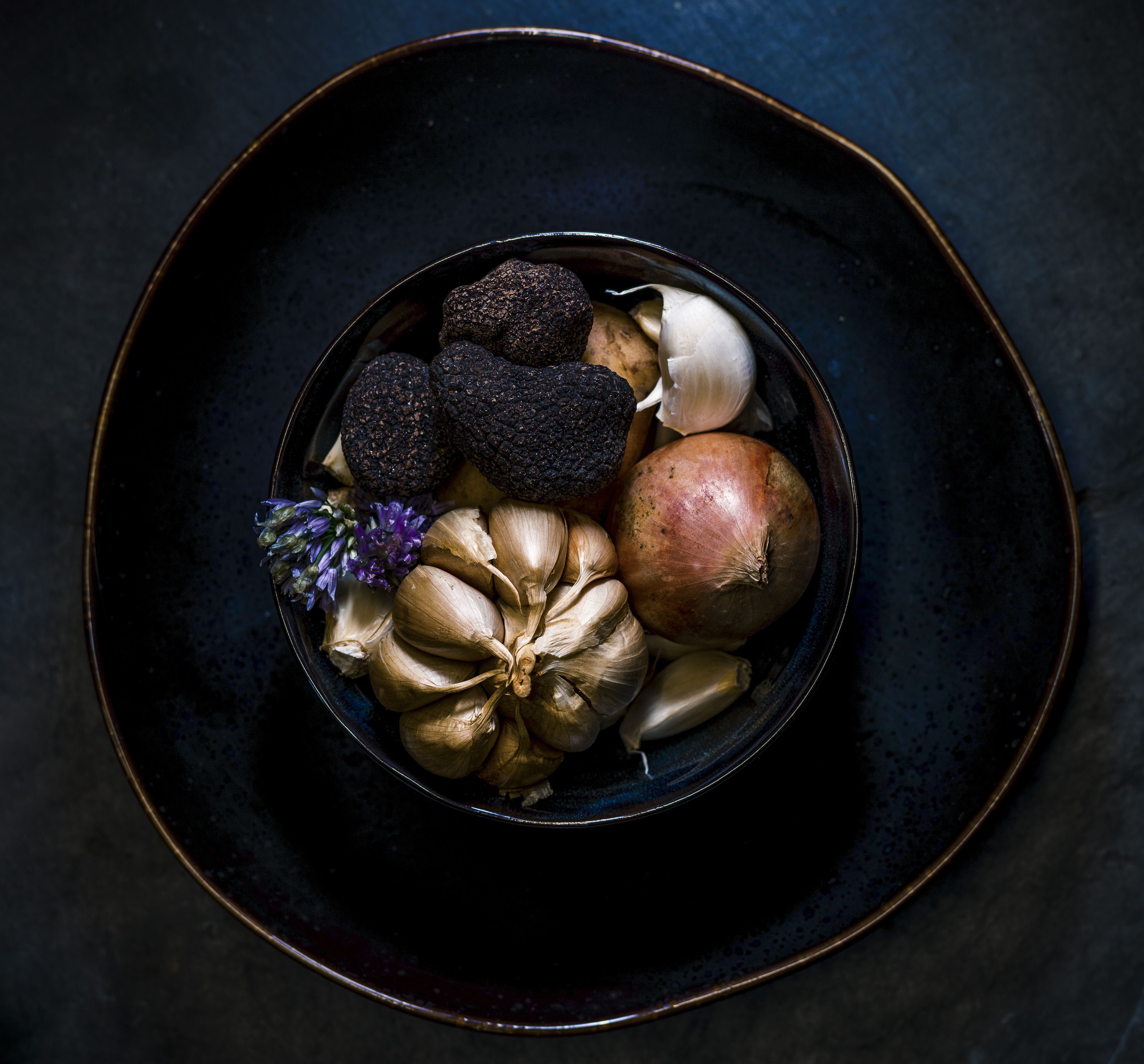 A bowl of assorted vegetables including onions, garlic, and black truffles on a dark plate.