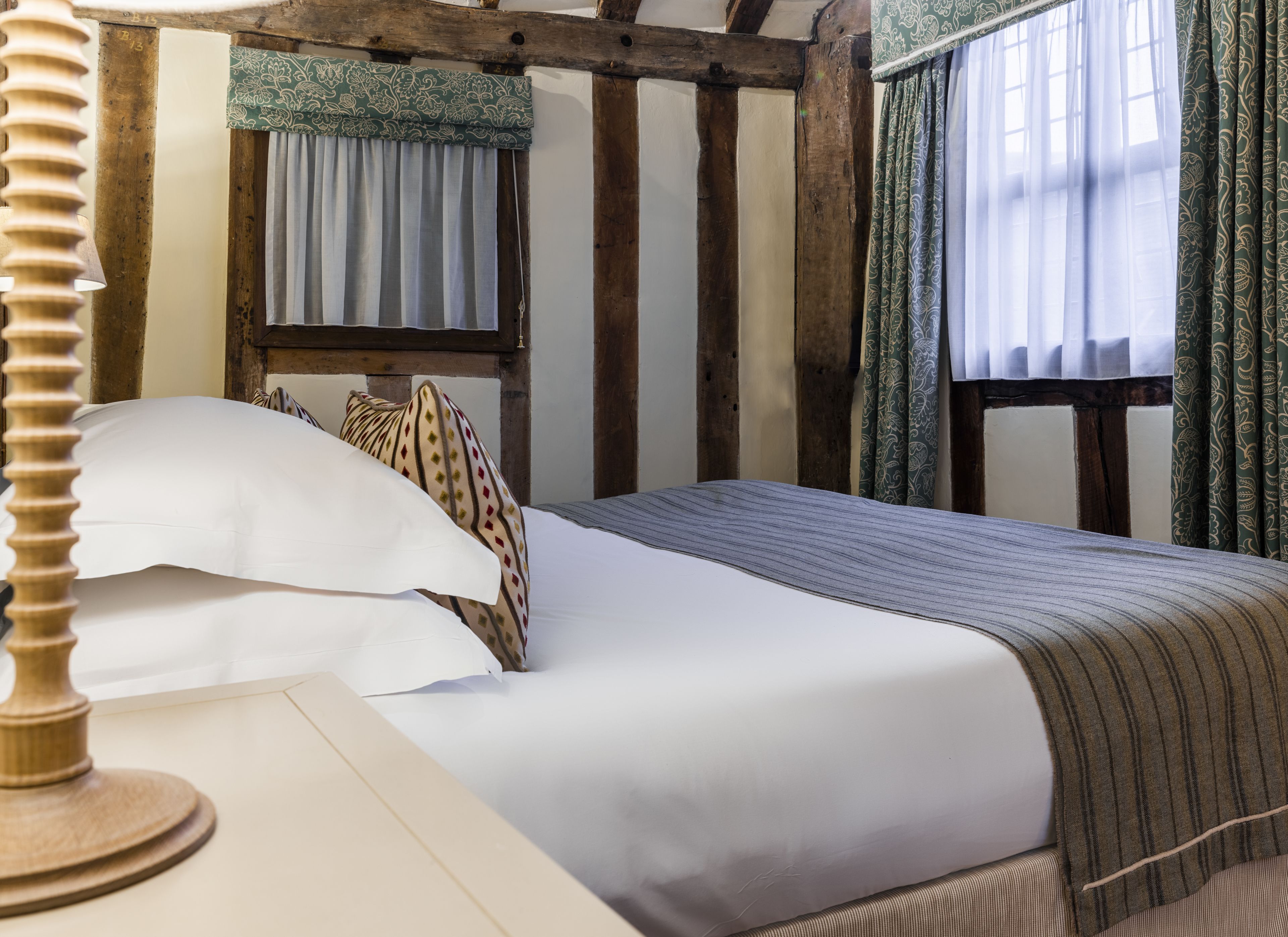A traditional room with exposed wooden beams, a bed with white linens, patterned pillows, and curtains with floral designs.