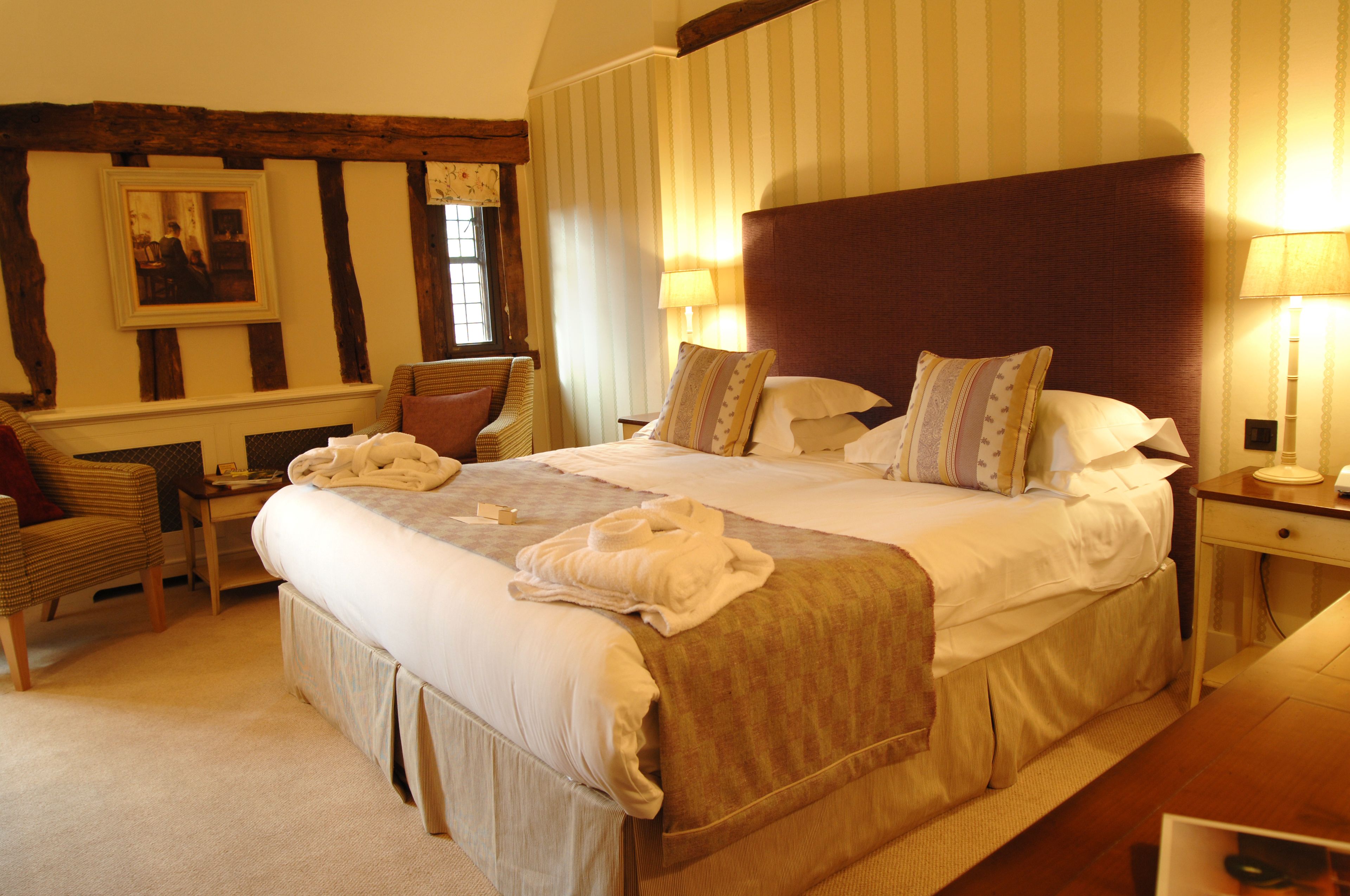 A large bed with a wooden headboard, a bedside table with a lamp, a chair, and a framed picture on the wall.