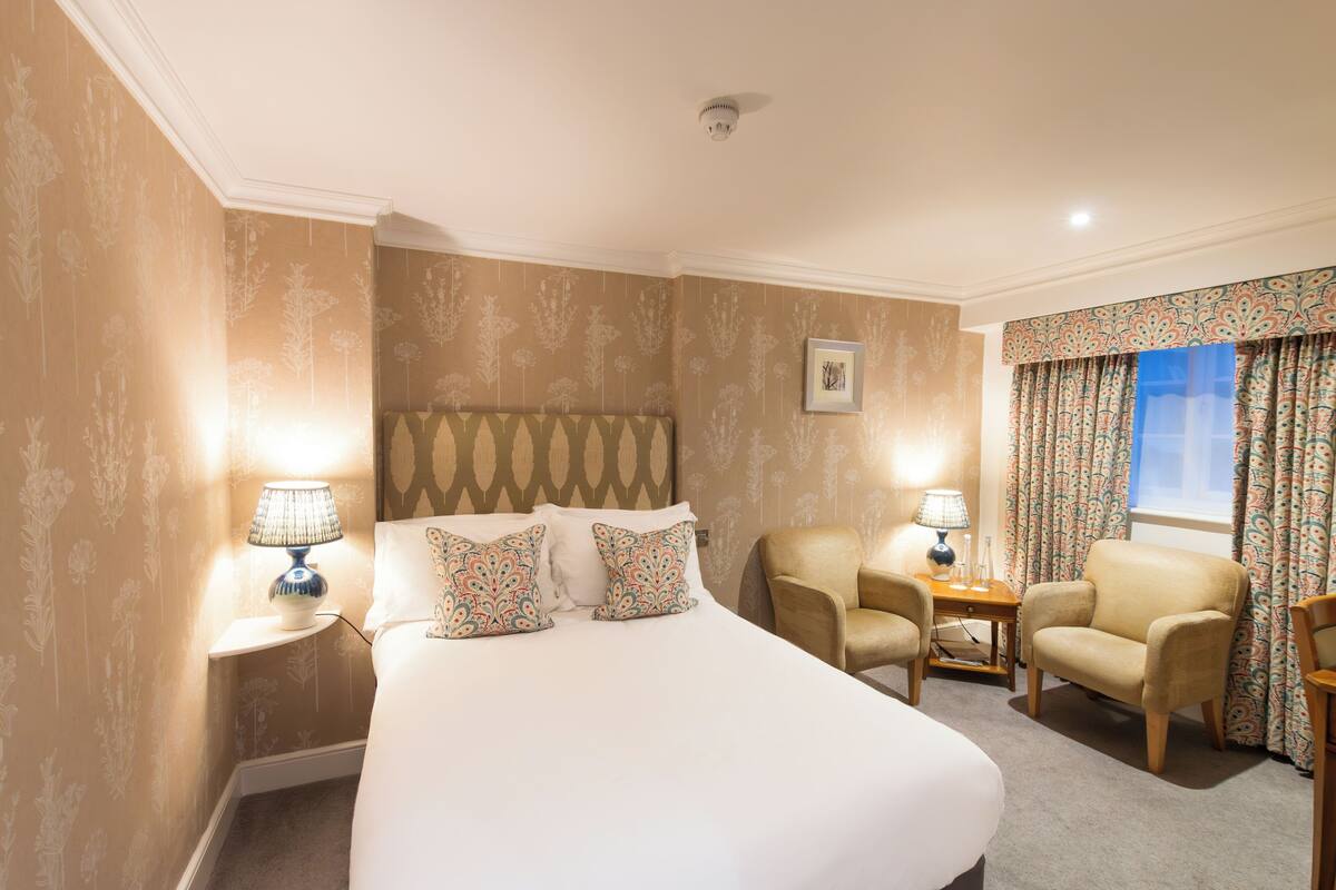 A hotel room with a large bed, two bedside tables with lamps, a chair, a small table, and a window with curtains.