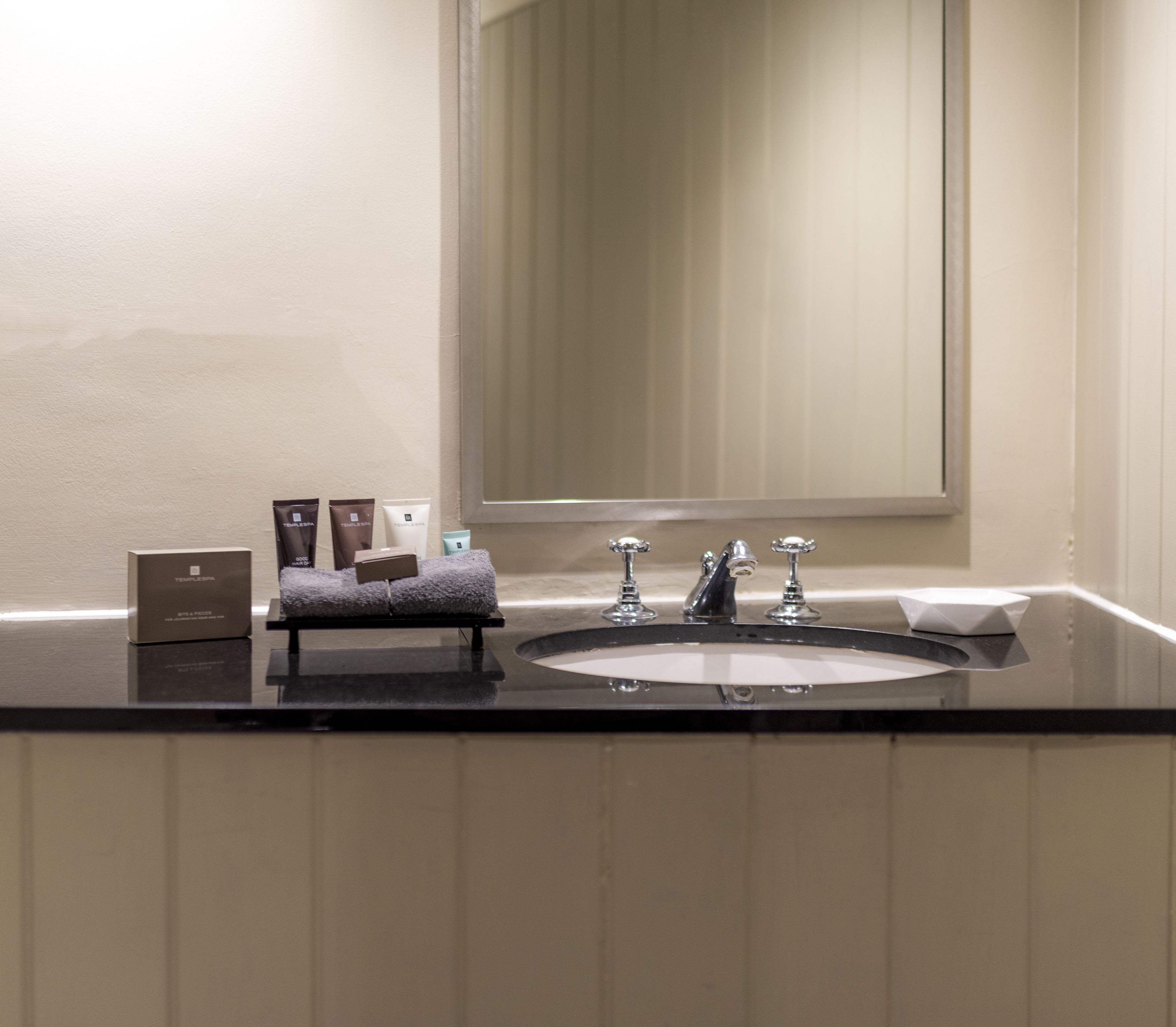 A bathroom with a large sink, two faucets, and a mirror above it. There are toiletries on the counter.