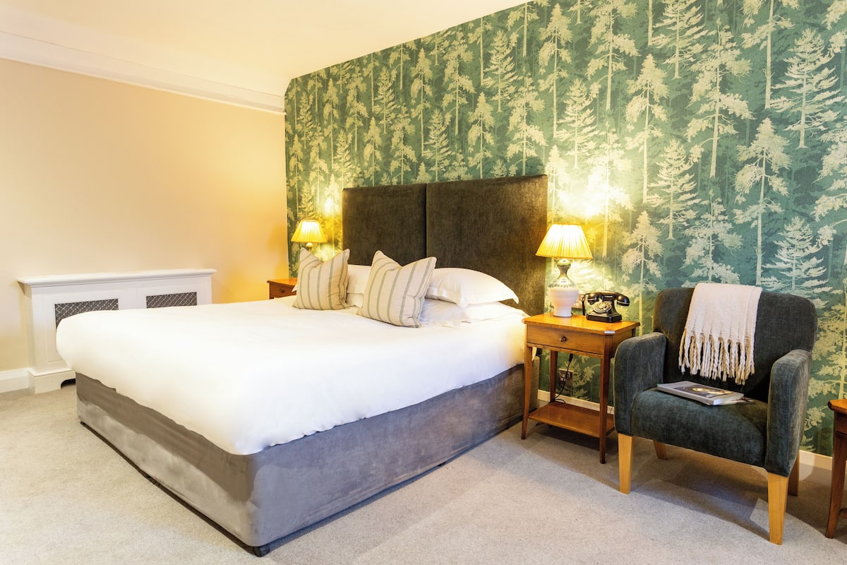 A bedroom with a large bed, two bedside tables with lamps, a chair, and a wallpapered headboard.