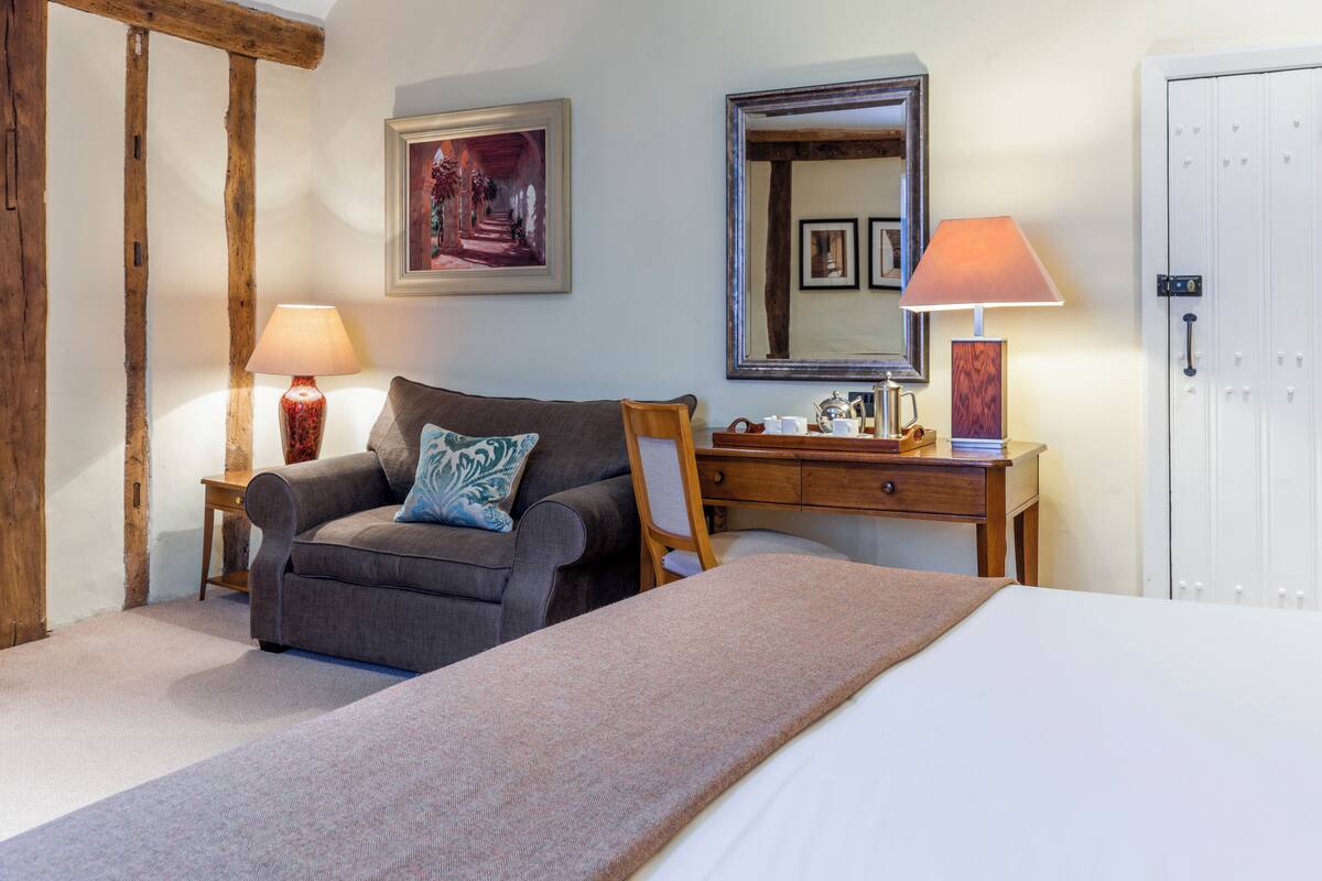 A bedroom with a bed, a sofa, a wooden desk, two lamps, a mirror, and a door.