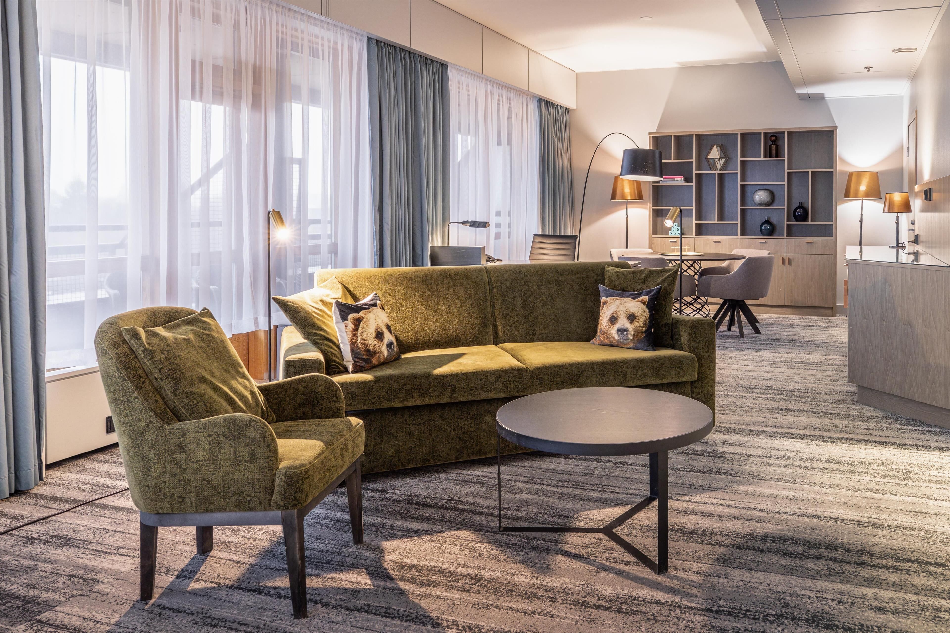 Una habitación de hotel moderna con un sofá verde, un sillón y una mesa de centro redonda. Hay un escritorio con una computadora portátil, una silla y una lámpara. La habitación tiene ventanas amplias con cortinas translúcidas y vistas a la ciudad.