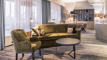 Una habitación de hotel moderna con un sofá verde, un sillón y una mesa de centro redonda. Hay un escritorio con una computadora portátil, una silla y una lámpara. La habitación tiene ventanas amplias con cortinas translúcidas y vistas a la ciudad.