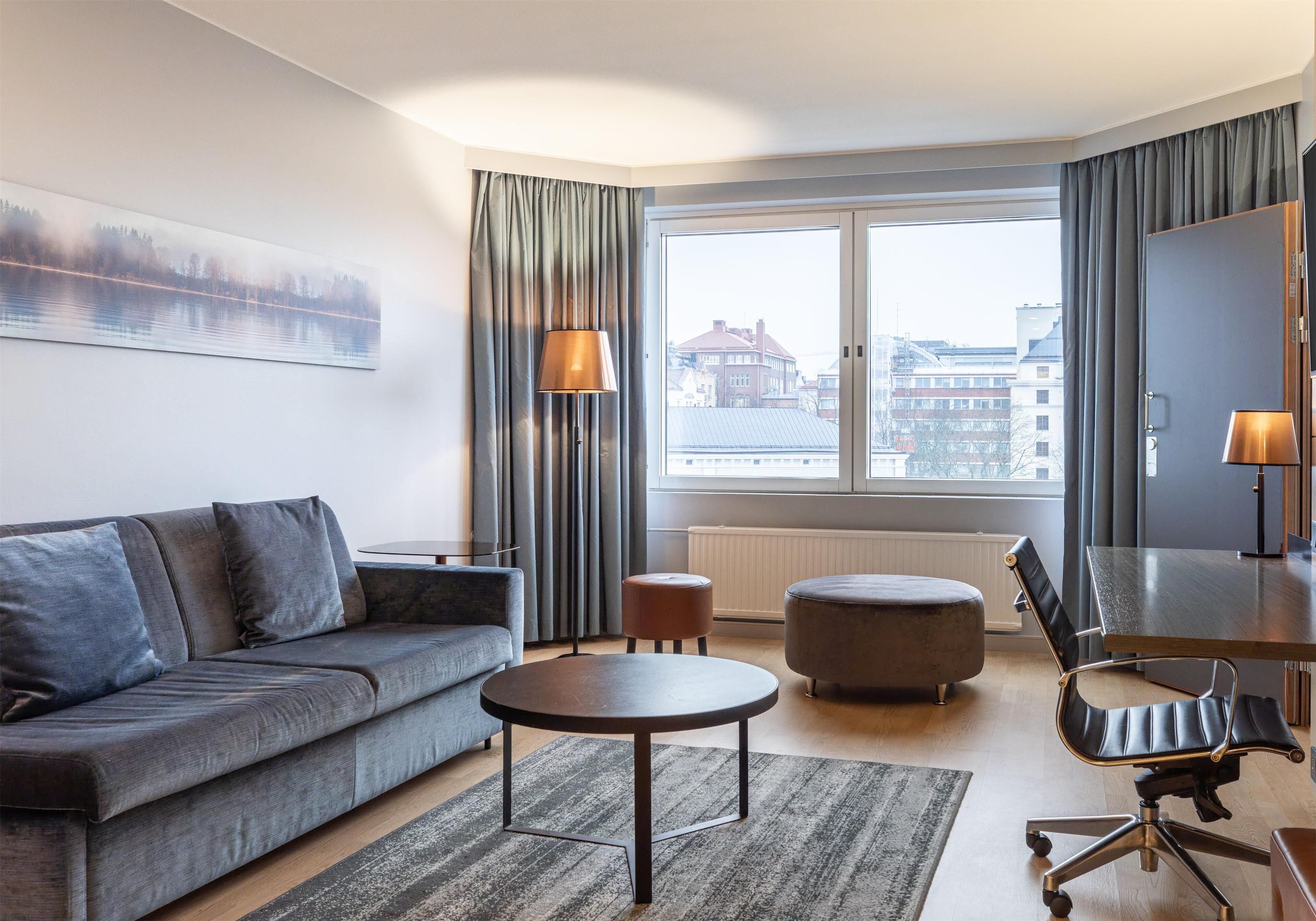 Una habitación de hotel moderna con un sofá gris, una mesita de centro redonda, una silla de oficina negra, un otoman pequeño y redondo, y un ventanal con cortinas.