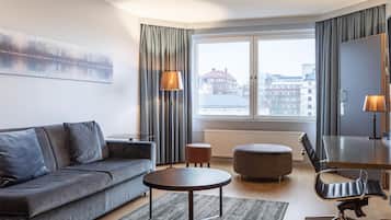 Una habitación de hotel moderna con un sofá gris, una mesita de centro redonda, una silla de oficina negra, un otoman pequeño y redondo, y un ventanal con cortinas.