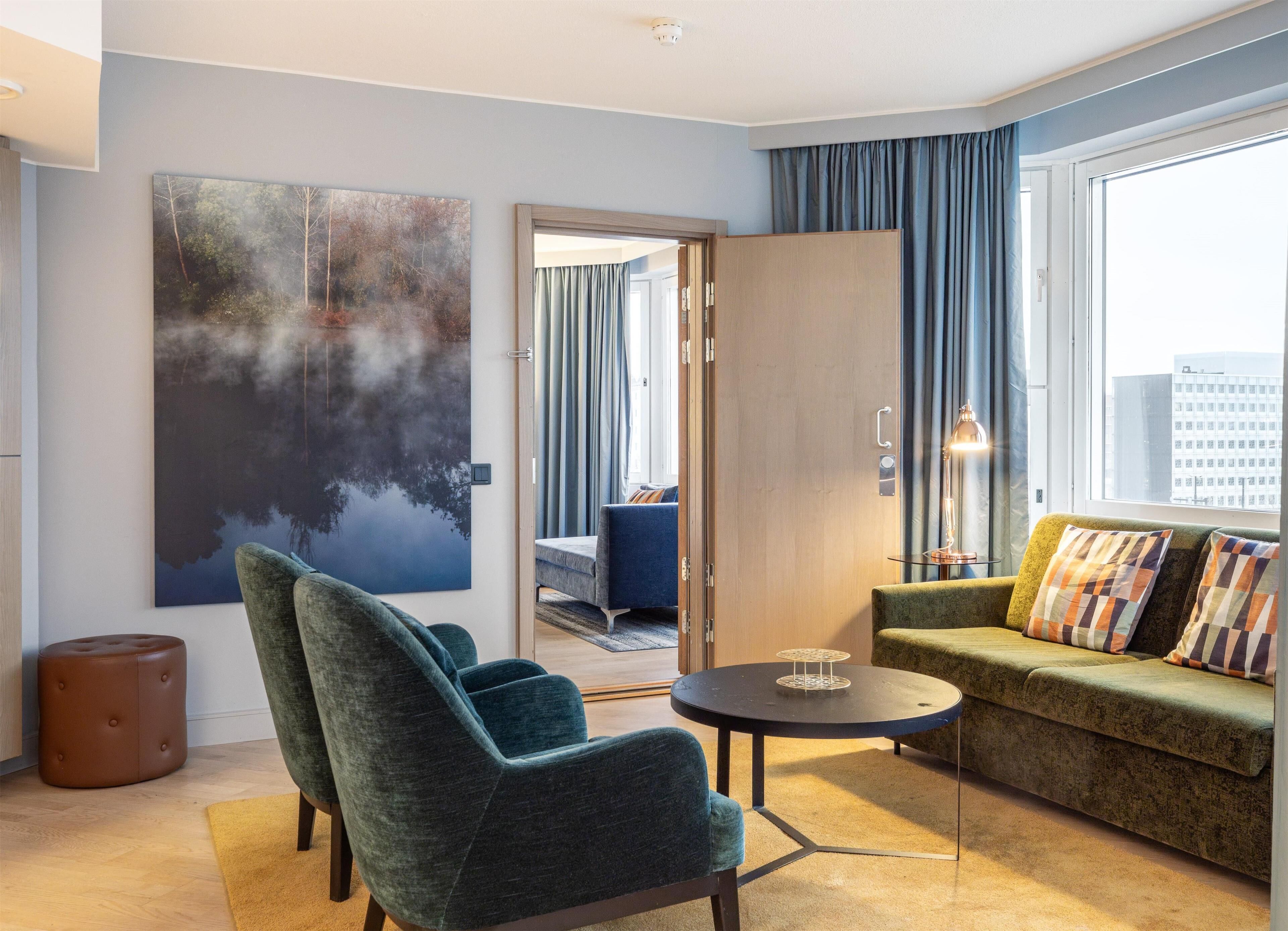 Moderni hotellihuone, jossa on suuri ikkuna, vihreä sohva, sininen nojatuoli, pyöreä pöytä ja suuri abstrakti maalaus seinällä.