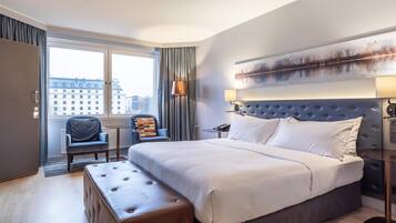 Habitación de hotel con una cama grande, una zona de estar con una silla, un escritorio y vistas a edificios a través de la ventana.