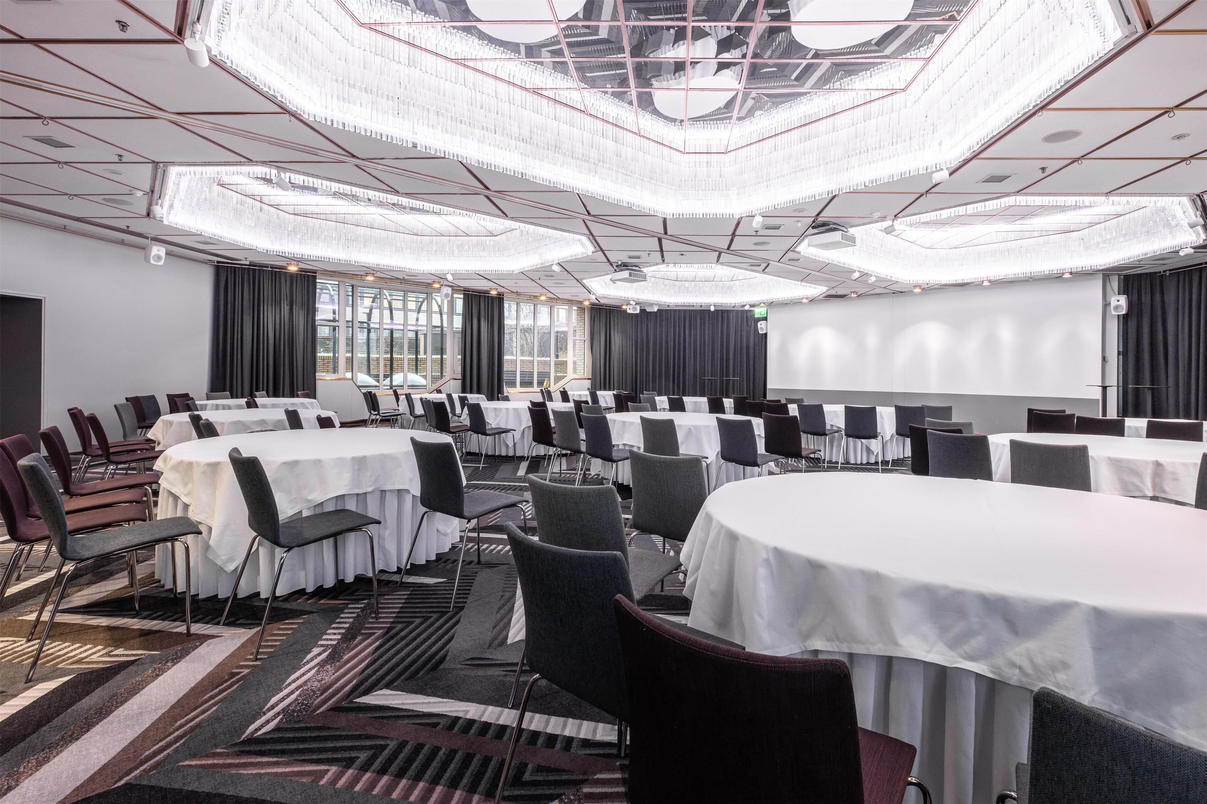 Un salón de conferencias con varias mesas y sillas, ventanas amplias y una lámpara moderna.
