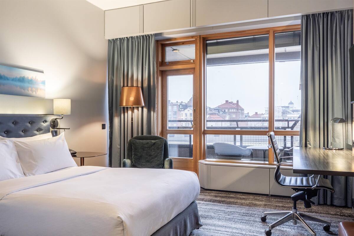 A hotel room with a large bed, a desk with a chair, a view of the city through a large window, and a painting on the wall.