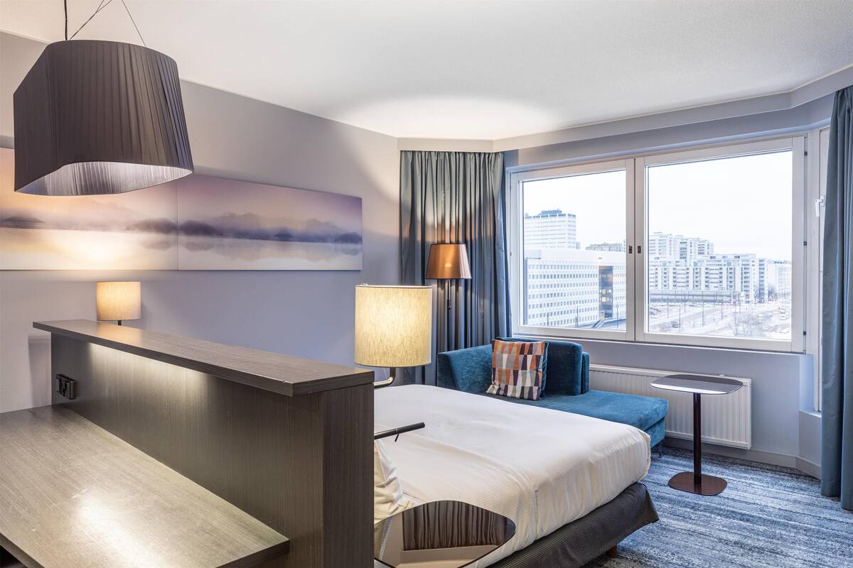 A modern hotel room with a bed, a desk, a lamp, a view of the city, and a large window with curtains.