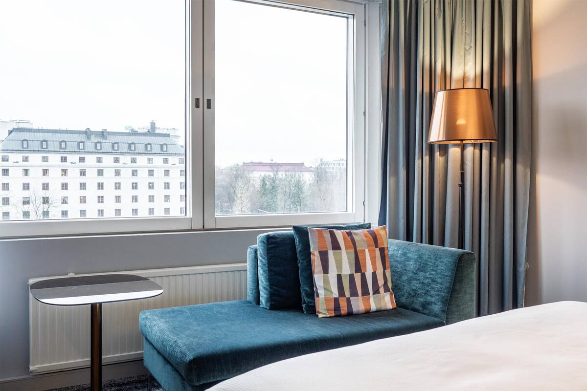 A hotel room with a bed, a small table, a lamp, and a view of buildings through a window.