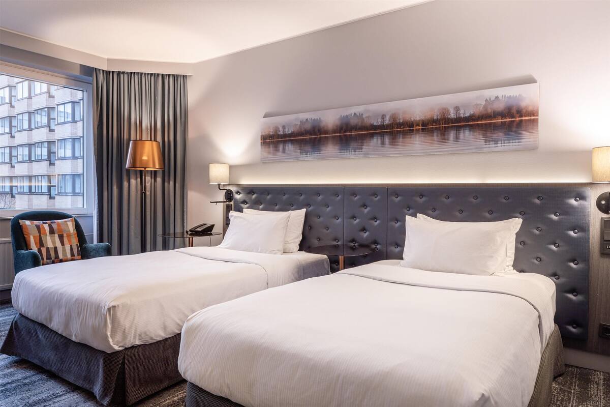 A hotel room with two beds, a large window with curtains, a wall-mounted painting, and a lamp.