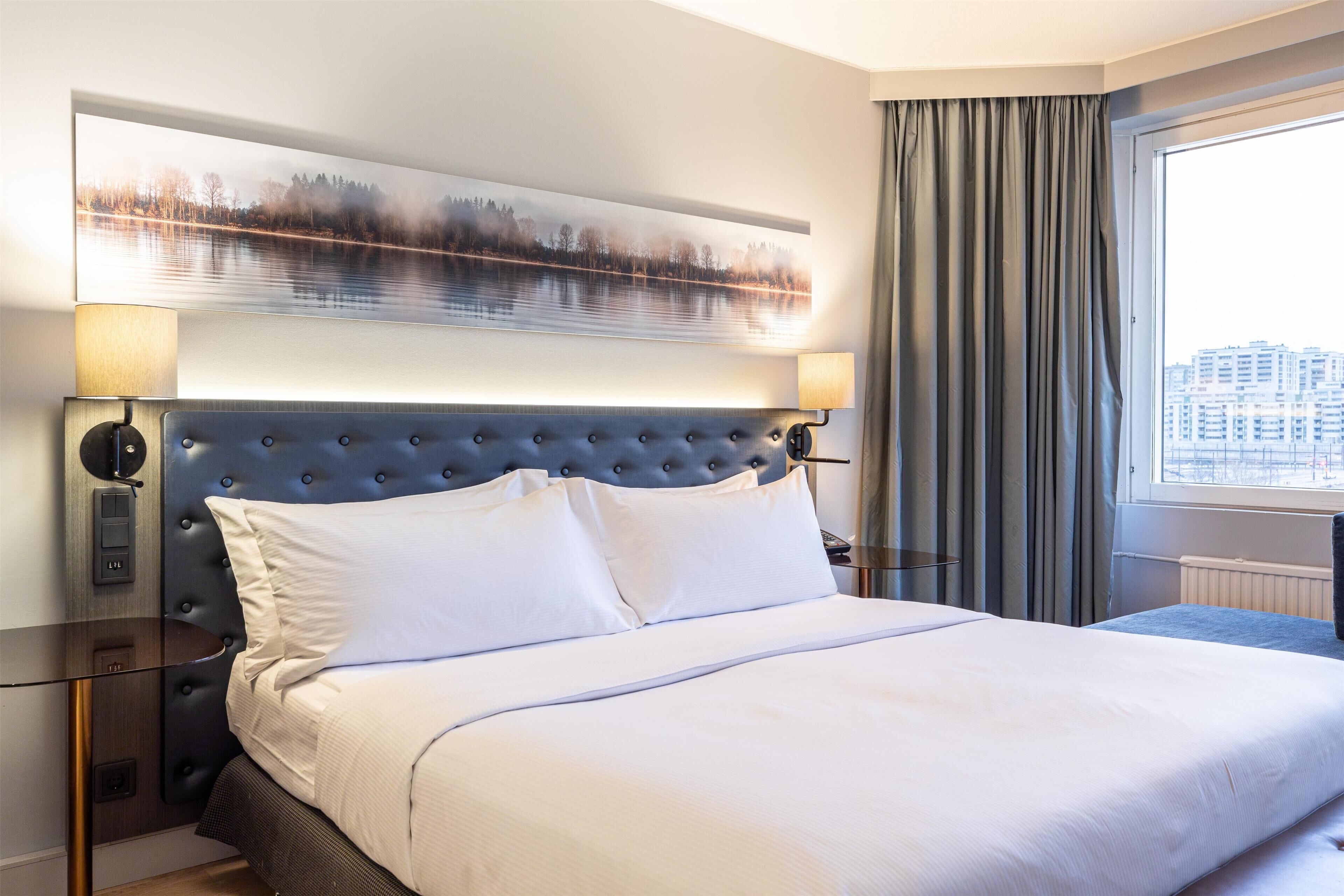 Ein Hotelzimmer mit einem großen Bett, einem gepolsterten Kopfteil, zwei Nachttischlampen, einem großen Fenster mit Vorhängen und Blick auf die Stadt.