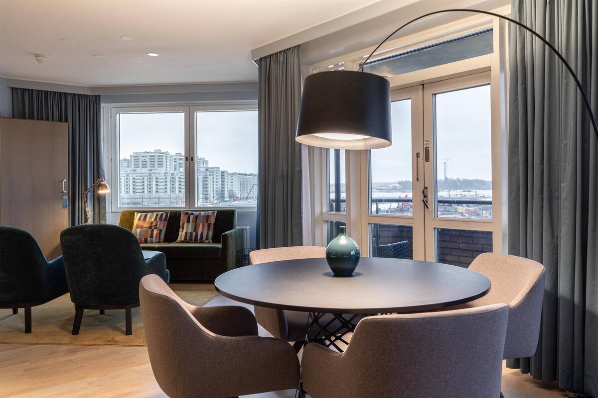 A modern living room with a round table, comfortable seating, a large window with a city view, and a floor lamp.