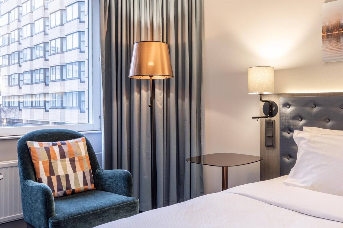 A hotel room with a bed, a chair with a patterned cushion, a floor lamp, a bedside table, and a view of a building through the window.
