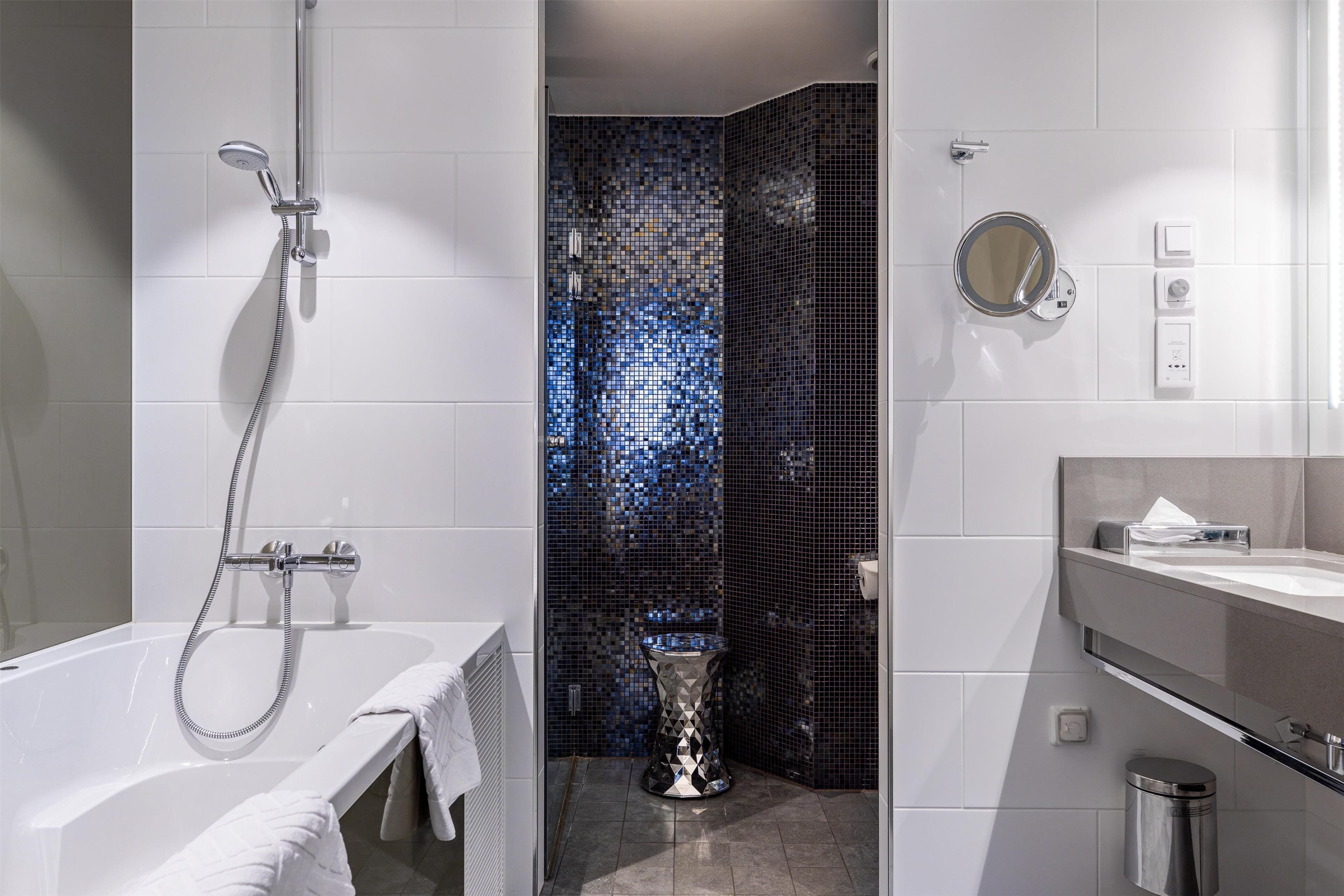 A modern bathroom with a white bathtub, a shower with a handheld hose, a toilet with a decorative base, and a vanity with a mirror and towel rack.