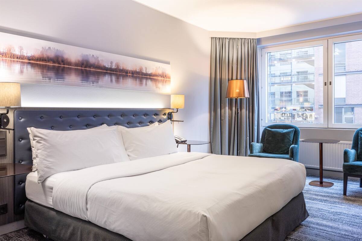 A hotel room with a large bed, a headboard with a landscape painting, a lamp, a chair, and a view of buildings through the window.