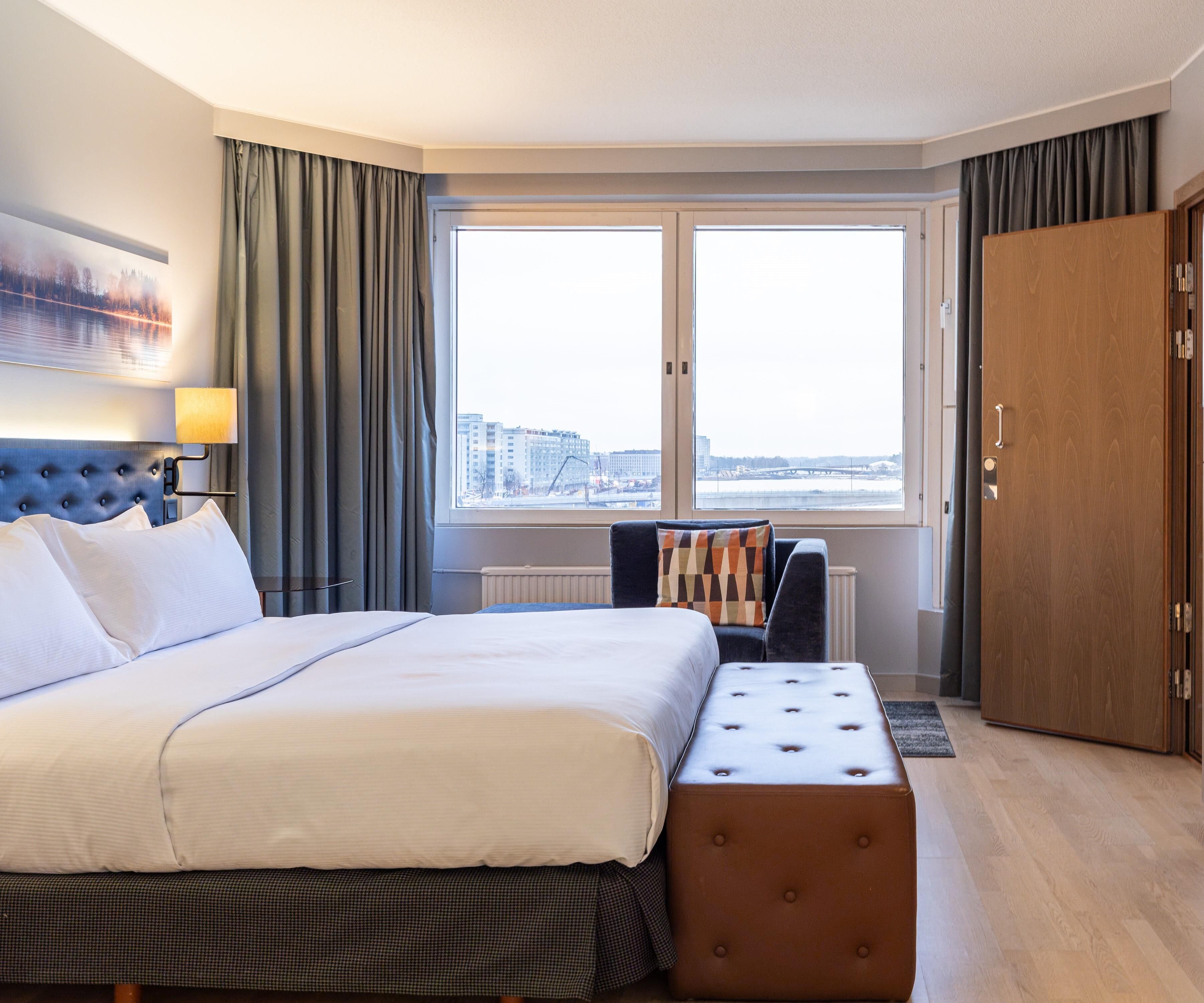 Habitación de hotel con una cama grande, un banco, vistas de la ciudad y un armario de madera.